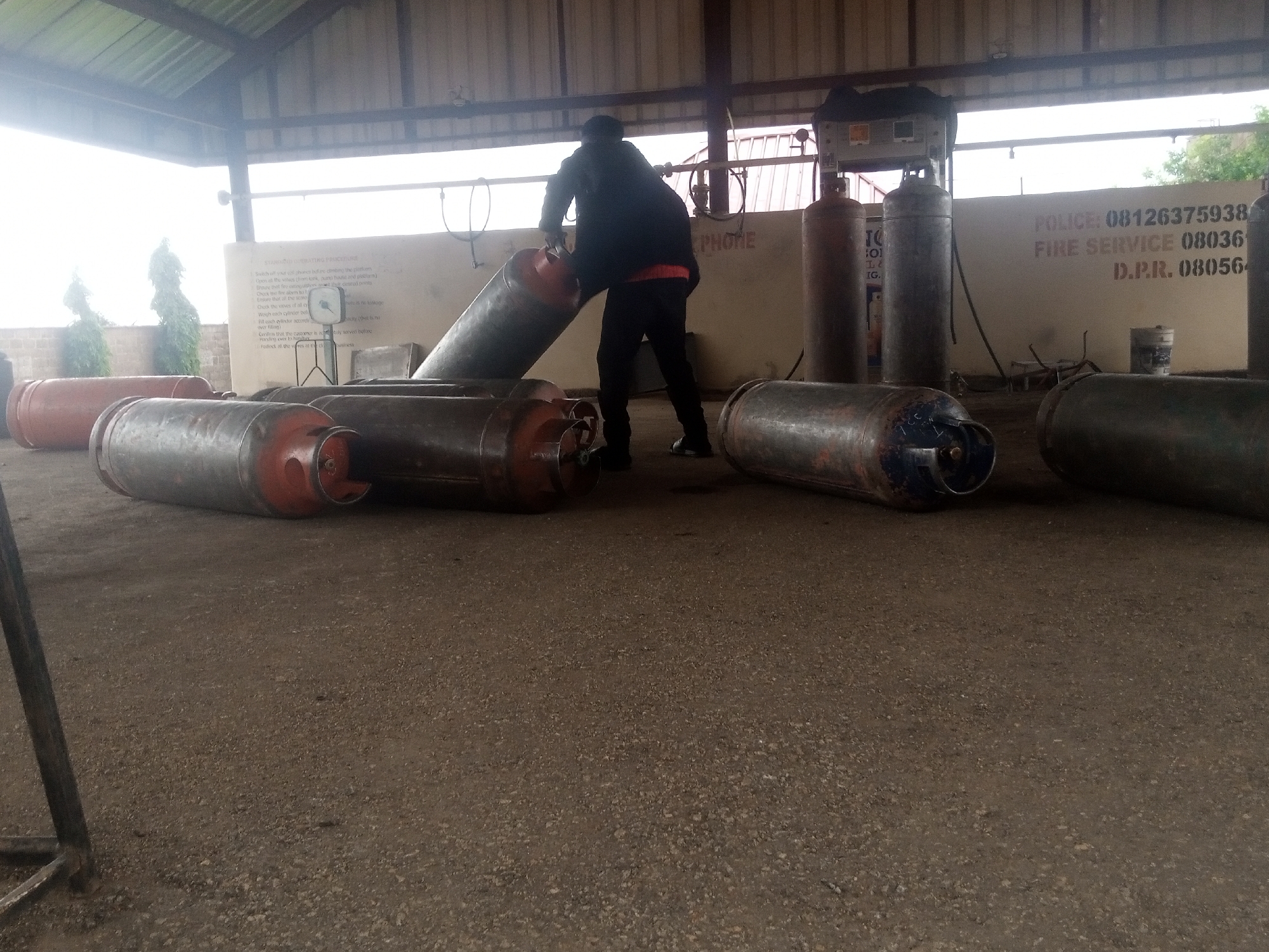 This media file shows a man in a dark jacket and pants handling a large, cylindrical object, likely a gas cylinder. He is in an open-air structure with a corrugated metal roof supported by metal beams. The ground is covered in gravel. To the left and right, several other similar cylinders are lying horizontally on the ground. In the background, a light-colored wall has text painted on it. The man appears to be engaged in a task involving moving or preparing the cylinders.
Visible text on the wall includes: "SAMICCI OPERATING TRICE", "PHONE", "POLICE: 08126375938", "FIRE SERVICE 08036", and "D.P.R. 080564". There is also a smaller sign with instructions. Two evergreen trees are visible through openings in the structure, suggesting an outdoor setting. The lighting suggests it might be daytime, possibly overcast or the structure is casting shadows. The scene appears to be a service area or storage facility for gas cylinders. The man's posture and action suggest he is actively working.
