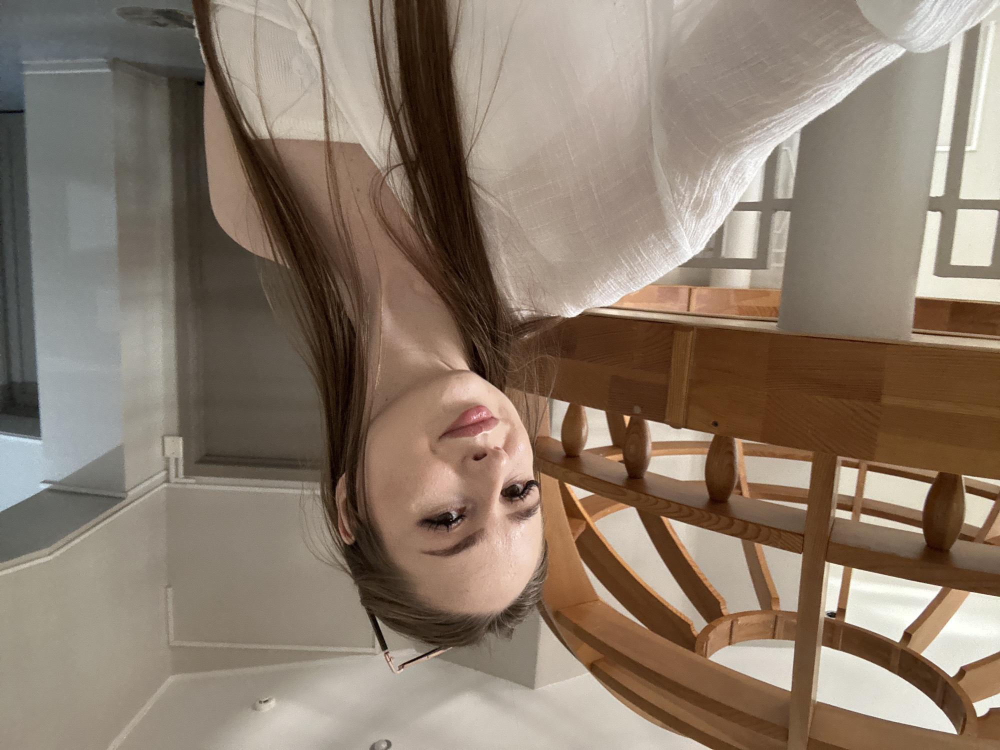 In an indoor environment, a young woman with long, brown hair and light skin is captured in a selfie. She is wearing a white top, and her gaze is directed towards the camera with a subtle smile. Her makeup includes mascara and lipstick. She is positioned in front of a wooden structure with a curved design. In the background, a gray wall and doorway are visible. The lighting in the scene is bright and appears to be from overhead sources.