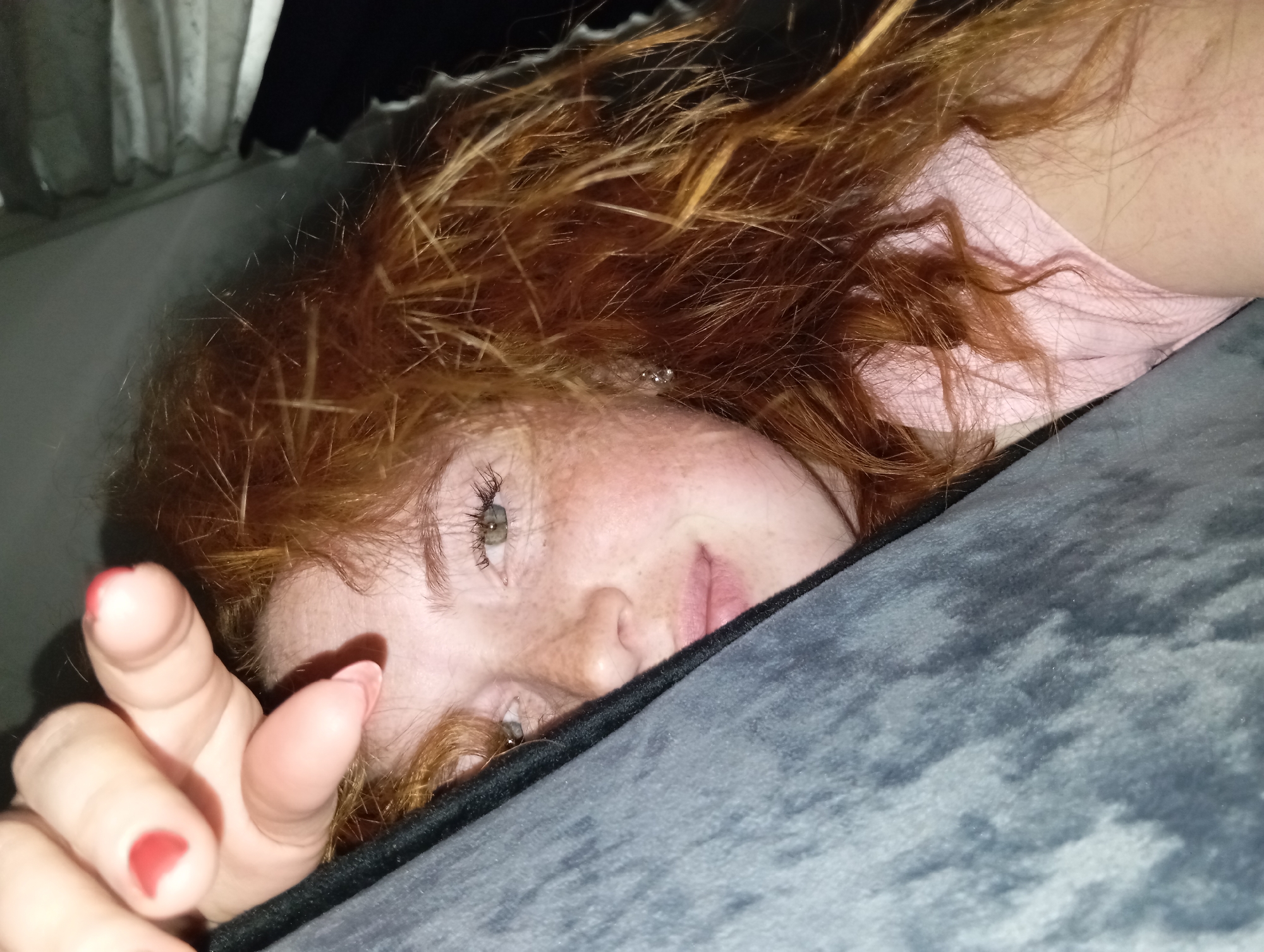 A close-up indoor shot focuses on a person lying down, partially covered by a dark grey patterned blanket. The person has red, curly hair and visible freckles on their face. Their eyes are open and appear to be a light color. A hand with red nail polish is visible in the foreground, with fingers extended towards the camera. A light pink garment can be seen under the hair and above the blanket. Behind the person is a dark space, possibly indicating a dimly lit room, with what looks like a curtain or window partially visible in the upper left corner. There is no information to confirm this picture was taken in the city of Yurgamysh, Russia.