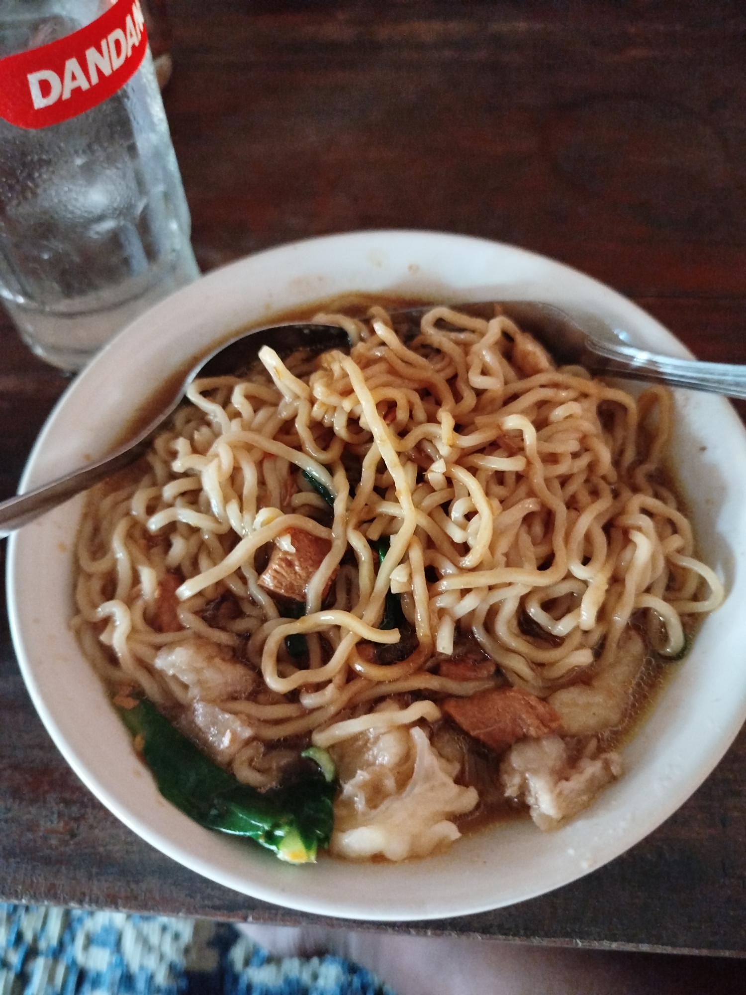 A close-up, overhead shot shows a bowl of noodles and a partially visible glass of water on a wooden table. The bowl is filled with golden-brown noodles, and pieces of meat and green vegetables are visible mixed in. A spoon rests on the edge of the bowl, and a fork is also present. The glass of water has a red label with white text that reads "DANDAN". The wooden table has a dark, grained surface. The background is a dark, out-of-focus wooden surface. The image appears to be taken in a casual dining setting, possibly a restaurant or home in Pojok, Indonesia.