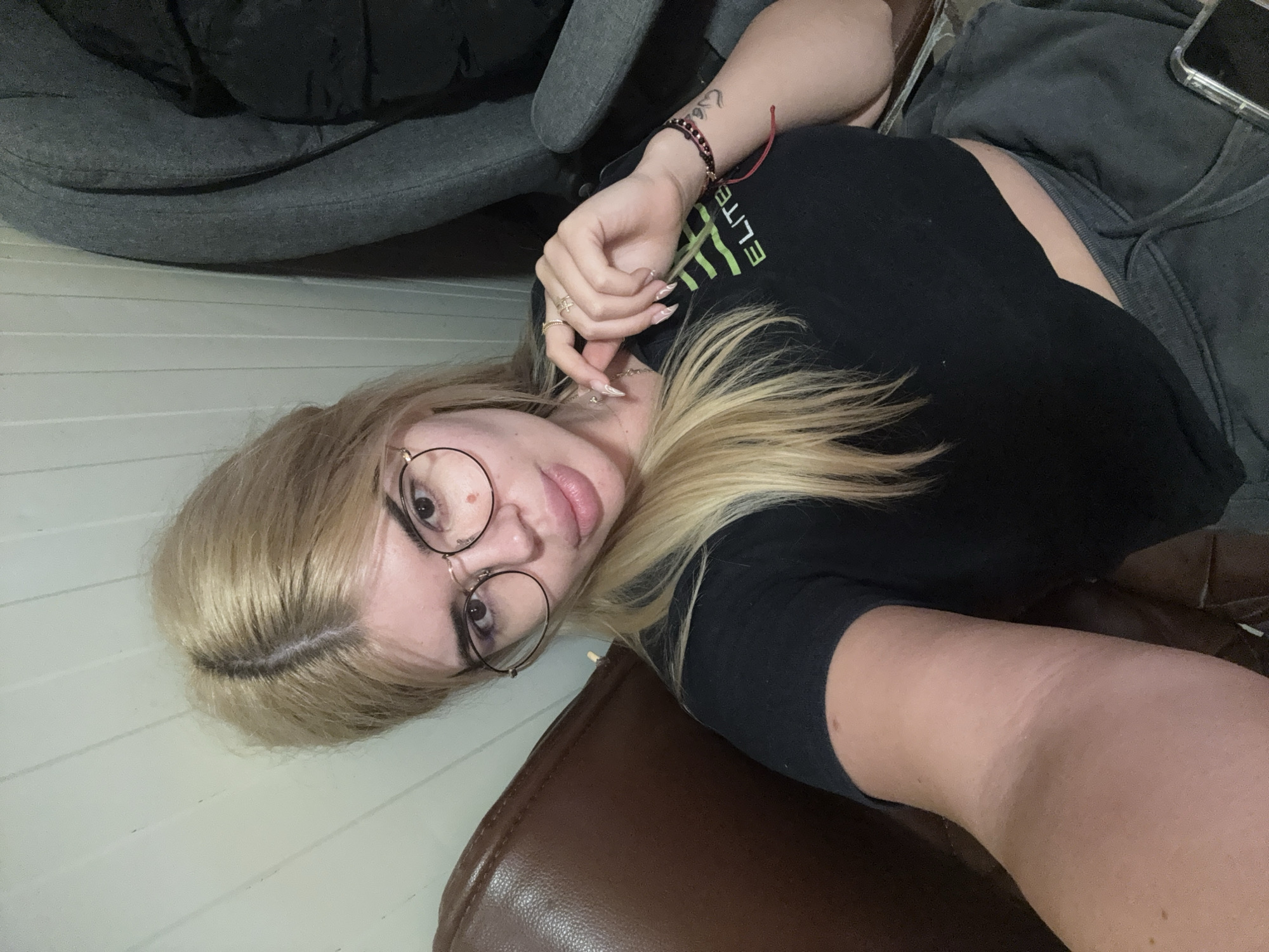 A young woman with blonde hair and glasses is lying down, facing the camera. She is wearing a black t-shirt and dark grey shorts. There is a tattoo on her right forearm and she is wearing several rings on her left hand. A red string is tied around her wrist. Her phone is visible on a dark grey blanket behind her. The scene appears to be indoors, with light-colored wooden paneling on the wall and a brown leather chair in the foreground. The lighting suggests it could be daytime or evening. There is no visible text in the image.