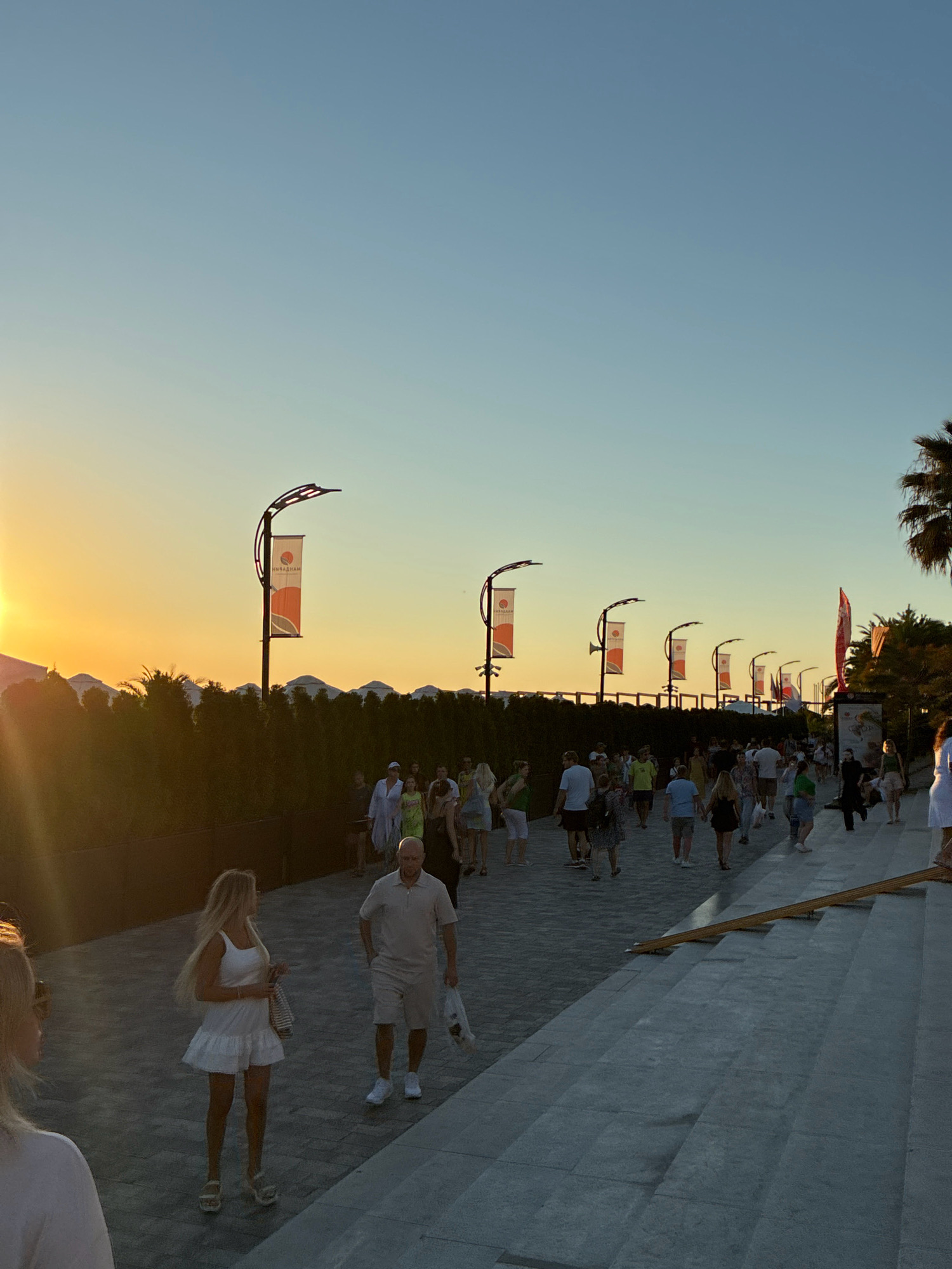 The image depicts a promenade in Sochi, Russia, during sunset. A diverse crowd of people walks along a paved pathway lined with decorative lampposts adorned with banners. The sun is setting on the left side of the frame, casting a warm glow and long shadows across the scene. In the background, lush greenery and palm trees can be seen. The atmosphere appears to be lively and relaxed, with individuals and groups enjoying an evening stroll.