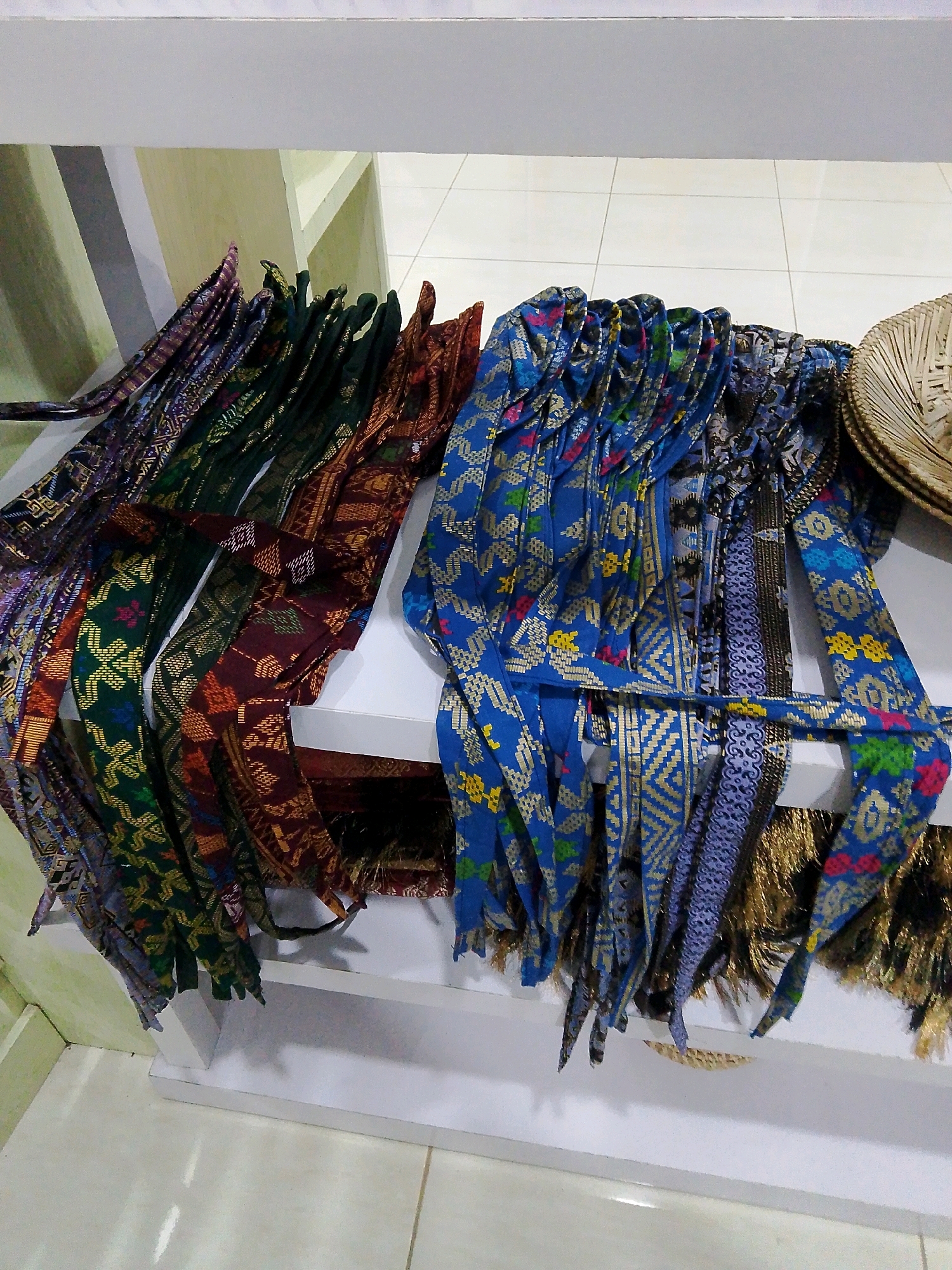 A horizontal, medium shot captures a display of intricately patterned woven textiles on a white shelf. The textiles are laid out in a pleated fashion, creating a vibrant cascade of colors and designs. Several distinct patterns are visible, including geometric motifs in blue with gold accents, and darker patterns in green and maroon with gold and red threads. To the far right, a portion of a woven basket is visible. The background consists of a white wall and a tiled floor. The lighting appears to be artificial, casting soft shadows. The scene is devoid of any people or animals, suggesting a retail or display setting. The overall impression is one of cultural artistry and craftsmanship. This image is likely located in Bunkate, Indonesia, a region known for its traditional weaving practices.