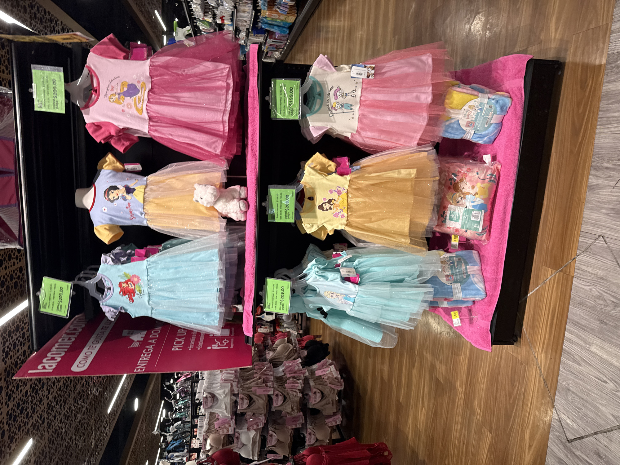 The image captures an interior retail scene within a La Comer supermarket, likely located in Puebla, Mexico. The primary focus is a two-tiered display of children's dresses.
The upper tier features three dresses on hangers: a light blue dress with Ariel from The Little Mermaid, a light blue and yellow dress with Snow White, and a pink dress with Rapunzel from Tangled. A small white, fluffy stuffed animal sits at the base of the Snow White dress. Each dress has a green price tag indicating a price of "$209.00." The lower tier also displays three dresses: a light blue dress with Elsa from Frozen, a yellow dress with Belle from Beauty and the Beast, and a pink dress featuring two cartoon characters and the text "Cutest in the nest." These dresses are priced at "$209.00" and "$199.00" respectively. The display stands are dark-colored, with each tier covered by a bright pink terrycloth-like fabric. Below the lower tier dresses, a pink package featuring Disney Princesses (likely a blanket or clothing set) and a bundled multi-colored "baby" item are visible.
In the left background, a large red promotional sign for "lacomer.com" is visible, displaying text in Spanish such as "COMO TE GUSTA TE LA LLEVES," "ENTREGA A DOMICILIO," and "PICK UP." Adjacent to this sign, shelves are stocked with numerous brassieres and lingerie items in various shades of nude and red. To the right background, additional retail shelving with various packaged goods is partially visible. The ceiling is dark and textured, illuminated by linear fluorescent light fixtures. The flooring is composed of light brown wood-look laminate planks, with a diagonal metallic strip or seam visible on the floor in the immediate foreground. No individuals are present in the scene.