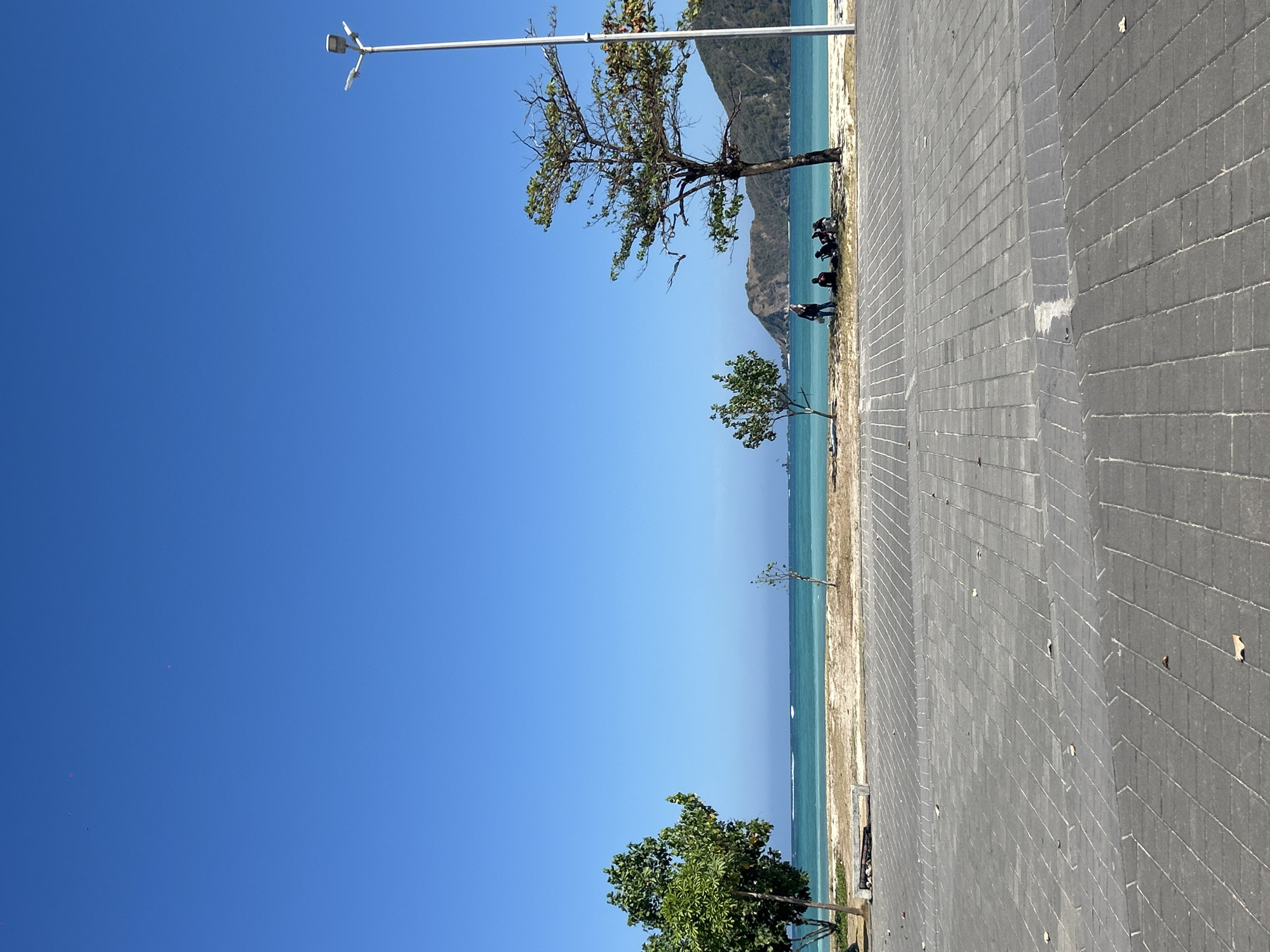 The image depicts a scene at Kuta Beach in Kuta, Indonesia, under a clear blue sky. In the foreground, a paved walkway constructed with grey rectangular tiles stretches across the bottom of the frame. The walkway has a textured surface with some fallen leaves scattered on it.
Beyond the walkway, a strip of sand forms the beach, with a few sparse trees dotting the landscape. To the left, a larger, leafy green tree stands prominently. Further to the right, a slender tree with fewer leaves is visible. A bench made of concrete and wood is situated near the larger tree.
The turquoise water of the ocean extends to the horizon, with small white waves breaking near the shore. In the middle ground, a small group of five individuals is gathered under the shade of a tree. One person is standing, while the other four are seated.
In the background, rolling hills covered in vegetation rise from the coastline. A tall, modern street lamp with multiple lights is positioned on the right side of the frame, extending upwards into the sky. A white boat is visible in the distance on the water. The overall impression is a serene beachside setting on a bright, sunny day.