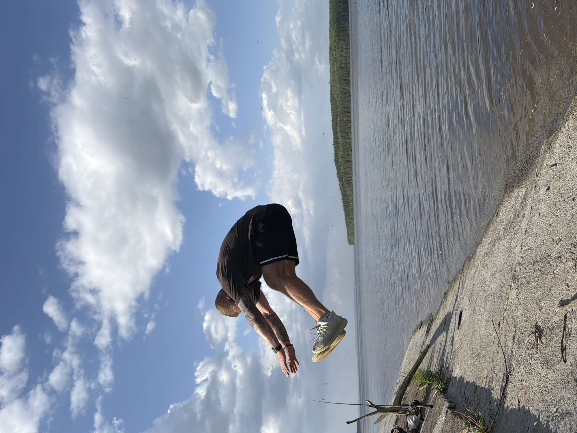 A man is performing a backflip near a body of water, likely a lake or river. The location is most likely in or near Chaikovsky, Russia. He is wearing a dark short-sleeved shirt, black athletic shorts with a white stripe, and light-colored athletic shoes. His arms are extended downwards toward the sky, indicating the peak of his backflip. He has tattoos on his arms.
The background shows a blue sky with white clouds. In the distance across the water is a green tree line. The water is a murky color. The ground near the water's edge is concrete. There is a fishing rod on the concrete area.