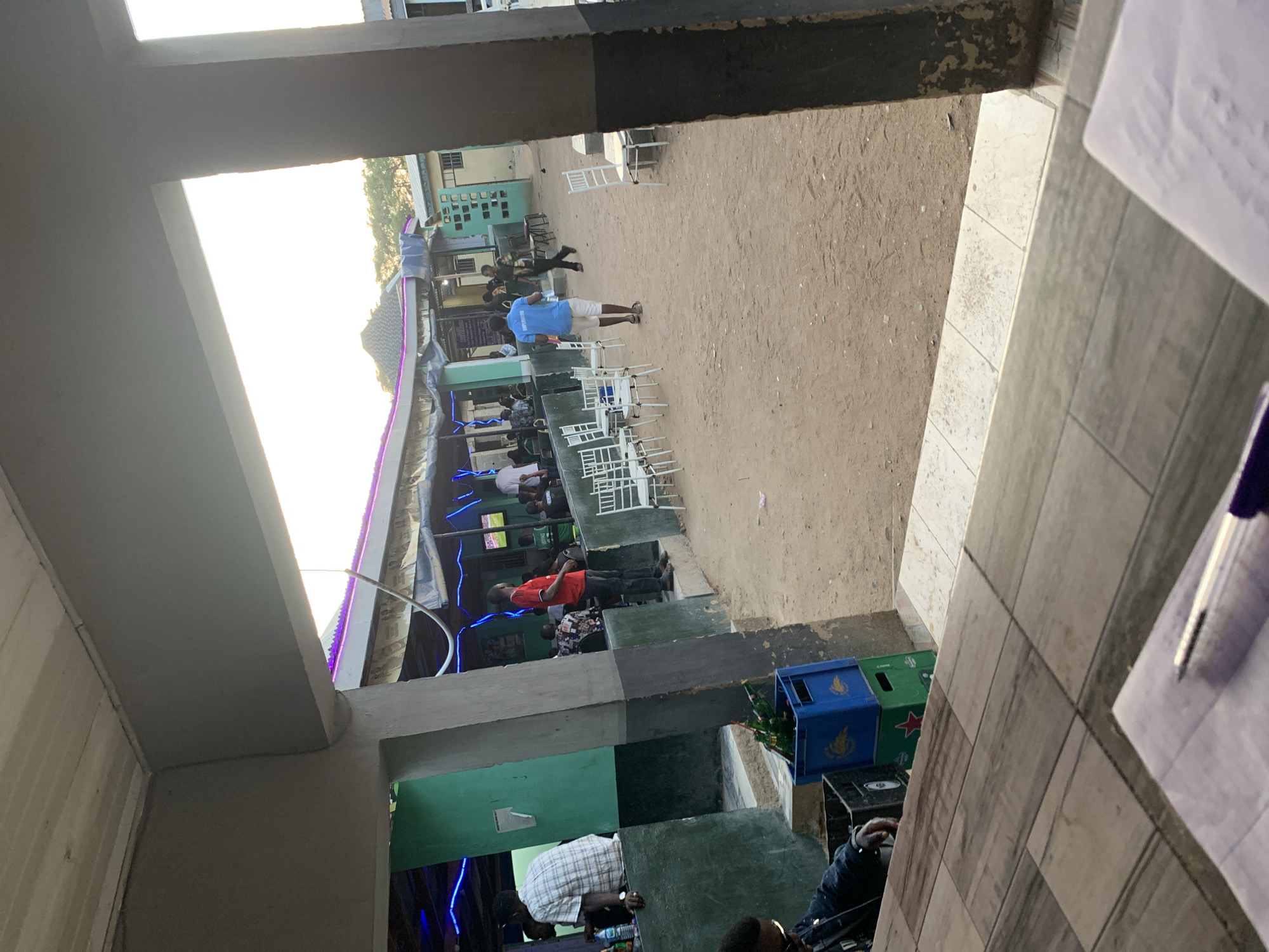 The image shows an outdoor event in New Stadium, Nigeria. People are seated at tables under a canopy adorned with blue lights. A man in a blue shirt stands in an open area. White plastic chairs are arranged nearby. In the foreground, a table with papers and a pen is visible. The scene is viewed from under a building's overhang, with concrete support structures visible on either side. Crates are stacked near the edge of the building. The ground is sandy. Buildings are visible in the background.