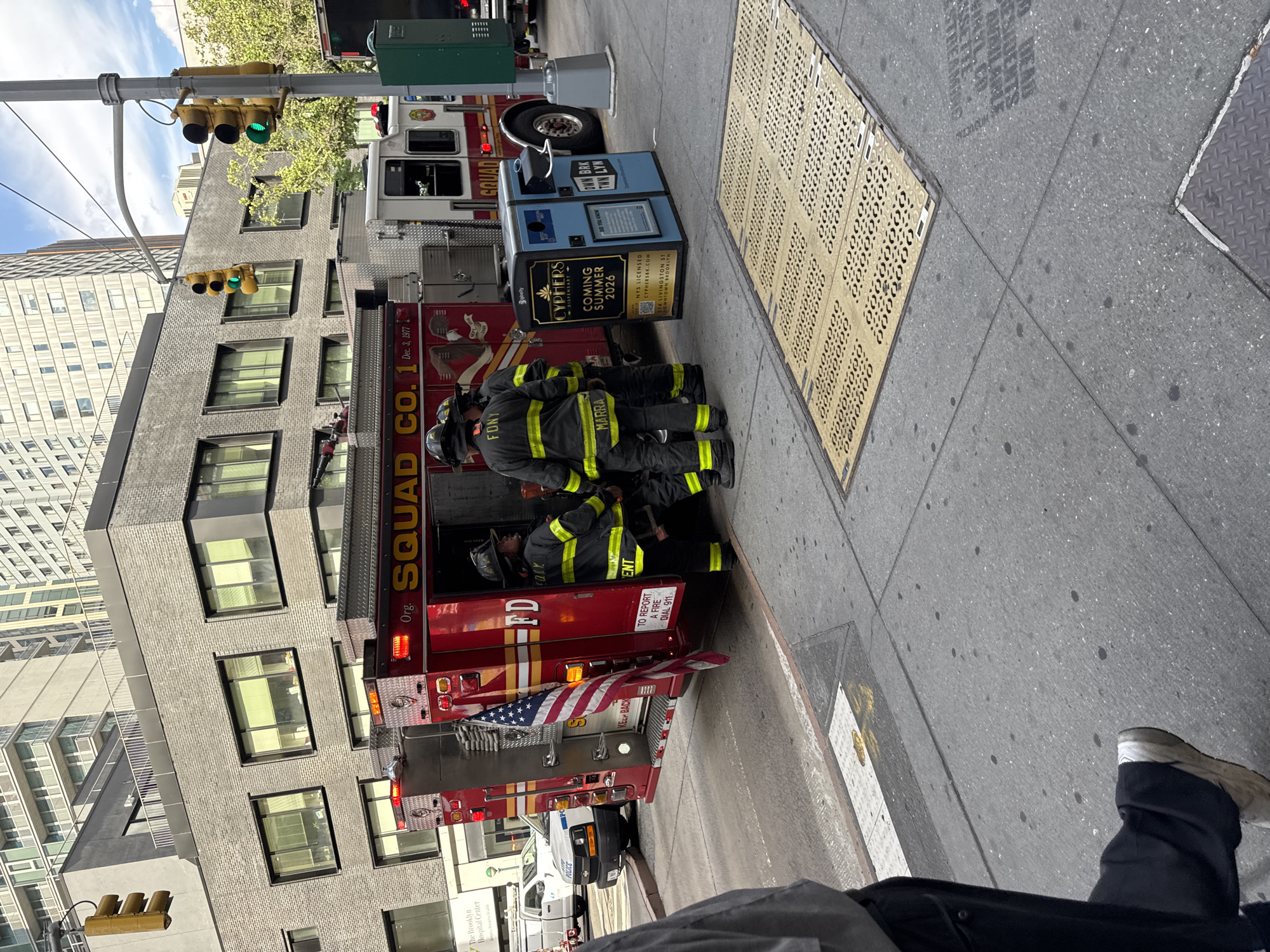 In this street-level scene in Brooklyn, New York, a red FDNY fire truck, marked "SQUAD CO. 1", is parked at an intersection. Three firefighters, dressed in full gear with helmets and reflective yellow stripes, are gathered near the rear of the truck. One firefighter, whose name tag reads "MARRA", is looking away from the camera. Another firefighter has "FONT" on their helmet. An American flag hangs from the side of the fire truck. To the right of the fire truck, a blue trash receptacle with advertisements for "CYPHERS DISPENSARY" and "COMING SUMMER 2026" is visible. In the background, modern buildings rise, one with a distinctive white facade and balconies. Traffic lights displaying green indicate the intersection is open. A police vehicle is parked behind the fire truck. The sidewalk is made of concrete, with a patterned yellow grate in the foreground. A person, mostly out of frame, is walking on the sidewalk in the lower left corner of the image. The sky is partly cloudy.
