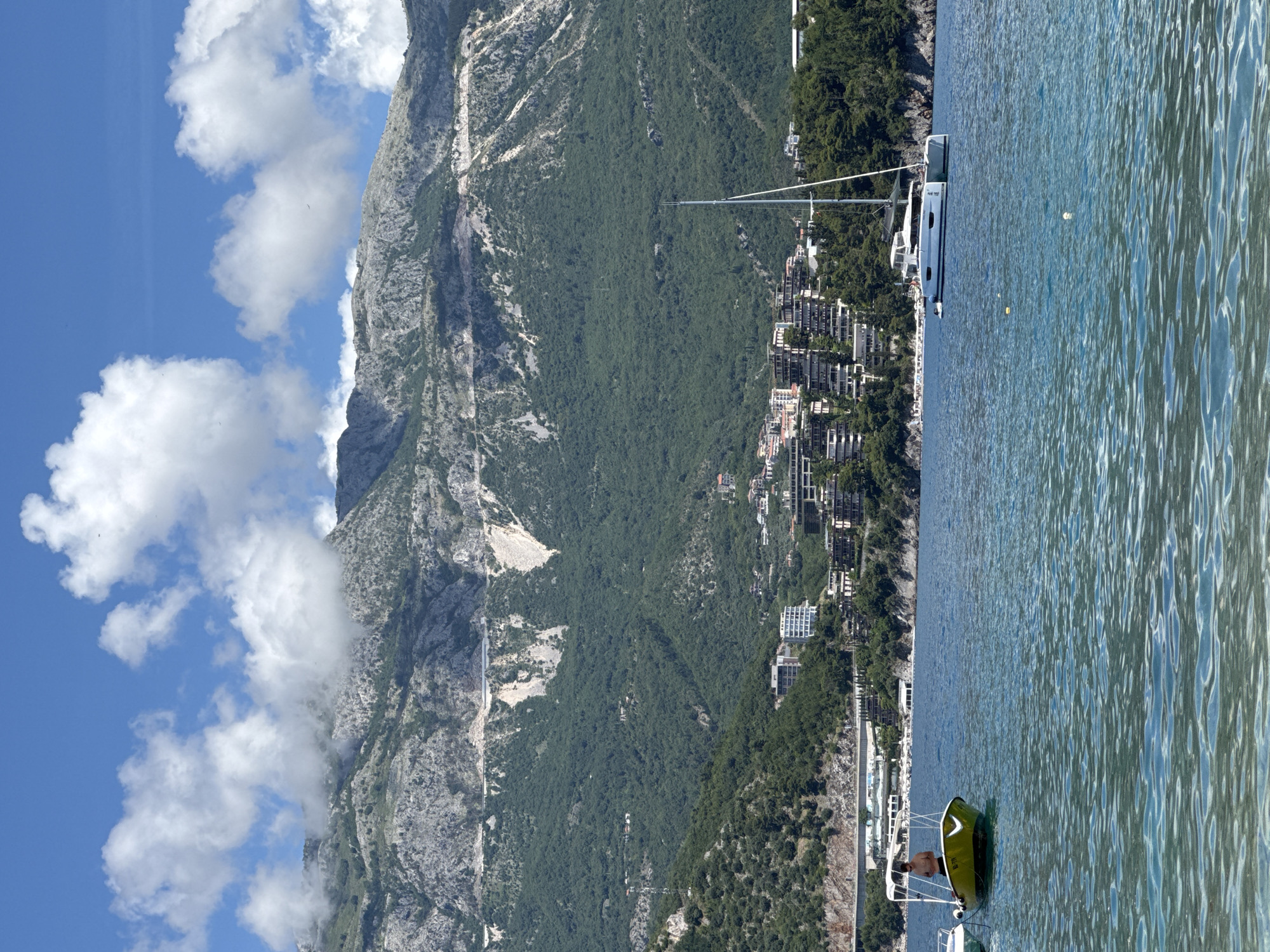 This image captures a panoramic view of a coastal landscape, likely Budva, Montenegro, under a bright, sunny sky. The scene is divided between majestic mountains, a developed coastline, and the expansive sea.
On the left side, rugged mountains rise steeply, characterized by grey rock formations and dense green vegetation. Some paths or roads are visible winding up the mountainsides. Fluffy white clouds drift across the blue sky above the peaks. Along the base of these mountains, a town or settlement is built into the landscape, featuring numerous multi-story buildings and structures extending towards the water's edge.
The right side of the image showcases the calm, blue-green waters of the sea, with gentle ripples reflecting the sunlight. Several boats are present. A large, white catamaran or sailboat is anchored or moored closer to the shore, its mast reaching towards the sky. In the foreground, a smaller, bright green boat is visible on the water, with a person standing or sitting inside. The alphanumeric text "XA-9" is clearly visible on the side of this green boat.
The time of day appears to be late morning or early afternoon, indicated by the strong sunlight and clear visibility. The weather is bright and pleasant, contributing to the tranquil atmosphere of this scenic coastal location.