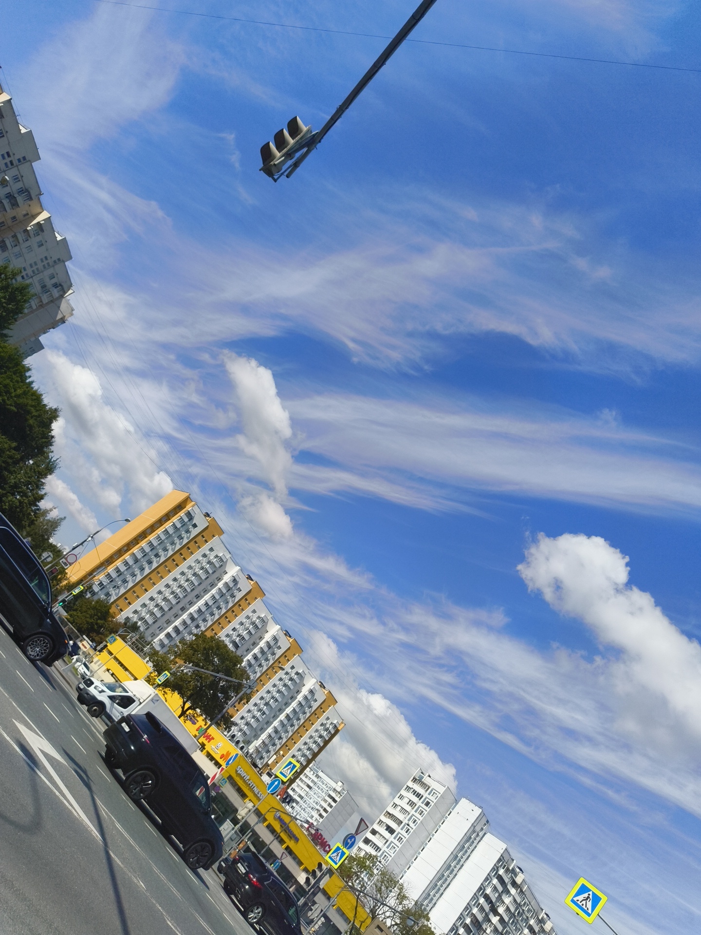 The image depicts a street scene in Moscow, Russia, characterized by a tilted perspective. A clear blue sky with wispy white clouds dominates the upper portion. A traffic light hangs from a wire spanning across the frame. On the left side, a tall, white residential building with multiple windows is visible, partially obscured by green trees. Further down the street, a yellow and white apartment building with numerous windows is prominently featured. Several cars are driving on the asphalt road, which has white lane markings. Two yellow pedestrian crossing signs with blue symbols are placed near the road. The overall scene suggests an urban environment on a sunny day.