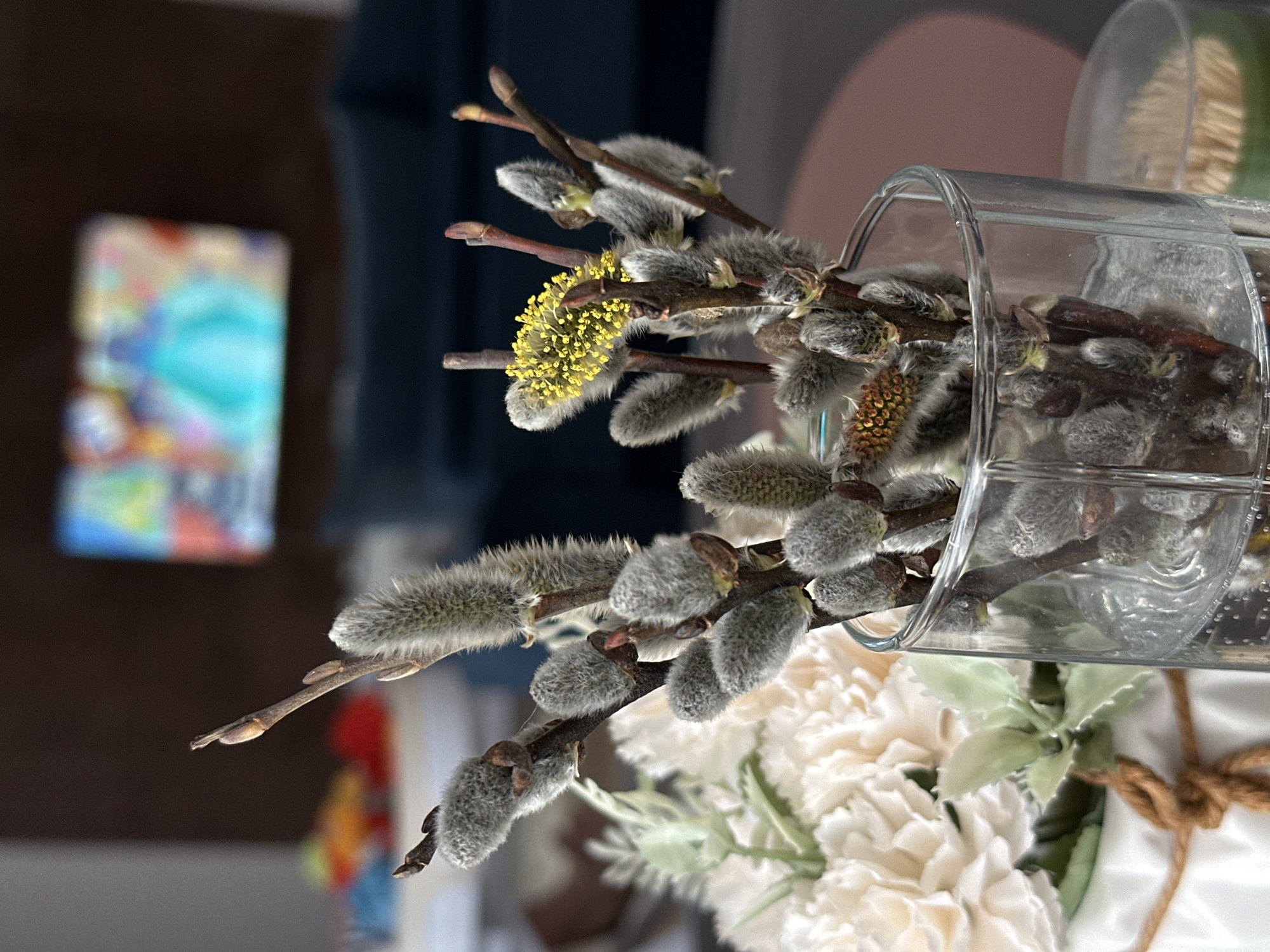 A still-life photograph shows pussy willow branches arranged in a clear glass vase. Some of the branches have begun to flower with yellow catkins. The vase is part of a larger floral arrangement that includes white flowers and green foliage. A section of the arrangement is wrapped with a coarse, natural fiber cord. In the background, a blurred object with a colorful, abstract design is visible. The environment appears to be indoors. There is no indication of the location being Подшивалово, Russia.