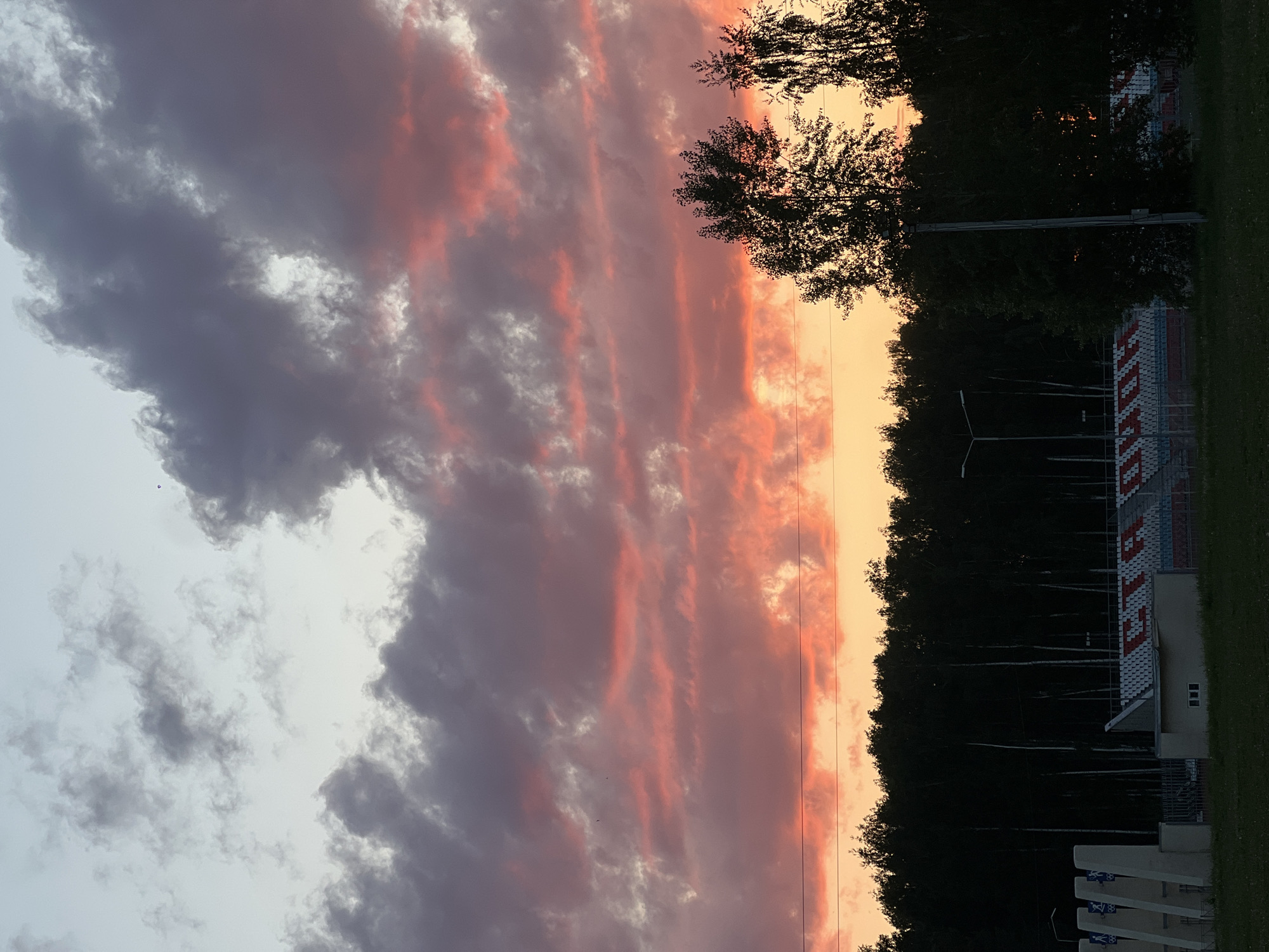The image depicts a stadium located in Chaikovsky, Russia, at twilight. The sky is dominated by long, horizontal clouds ranging from light gray to deep purple, illuminated with orange and pink hues from the setting sun. The right side of the image features a row of trees silhouetted against the sky. In the lower right, the stadium structure is visible, displaying seating and support buildings, and decorative columns. The overall scene captures a blend of natural and man-made elements at dusk.