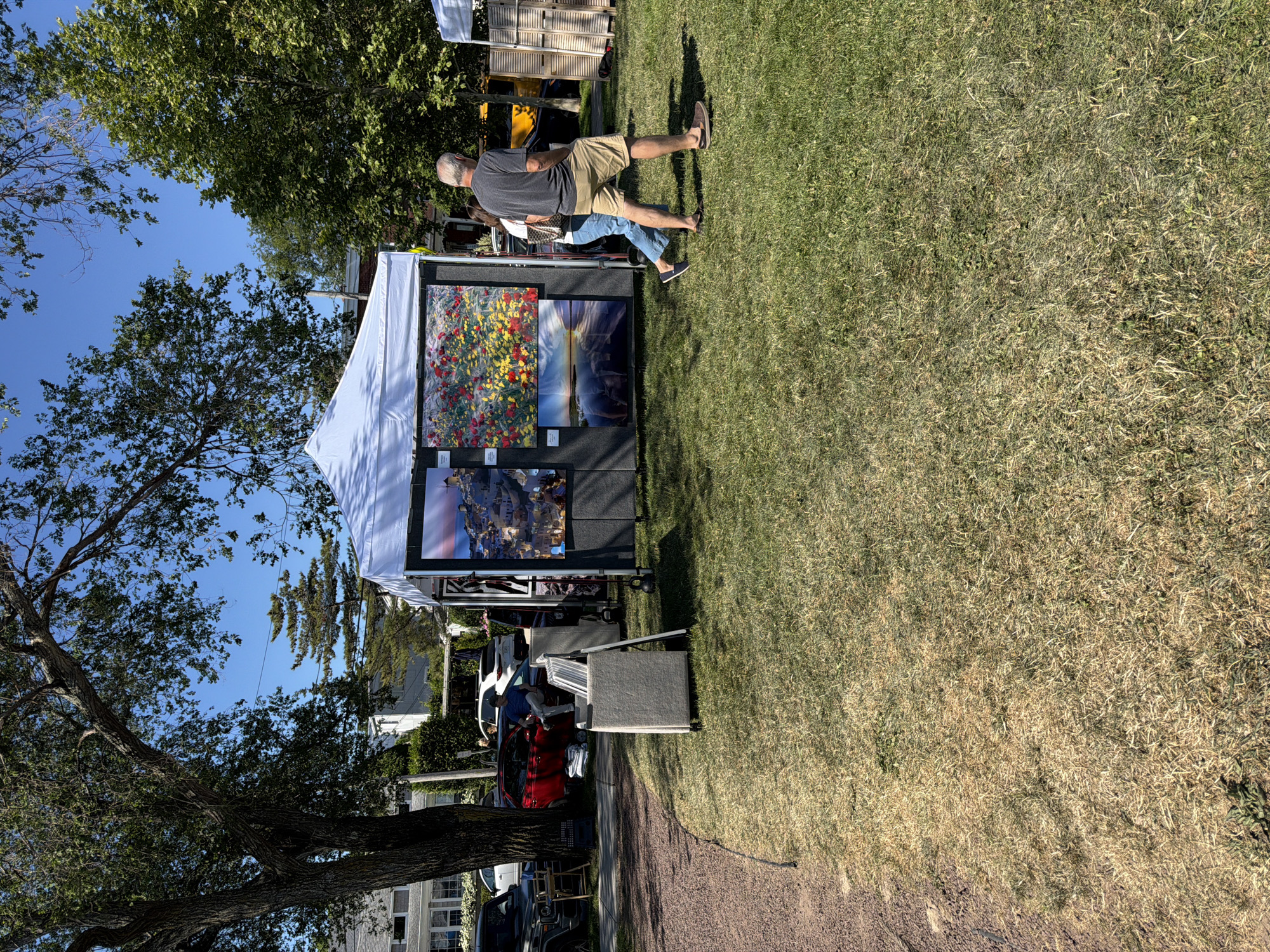 In the Village of Sag Harbor, United States of America, on a sunny day, a white tent displays art for sale. Two people, a man and a woman, walk past the art display. The man is wearing a dark t-shirt and khaki shorts, while the woman is wearing a white shirt and blue jeans. Behind the tent, vehicles are parked, including a red car and a black SUV. The art tent is set up on a grassy area adjacent to a gravel path. Trees with green leaves are visible in the background, and a clear blue sky completes the scene.