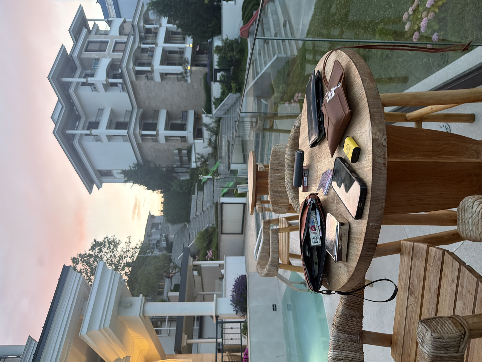 This image depicts an outdoor seating area in front of a modern apartment building, likely in the city of Obzor, Bulgaria, during sunset. A round wooden table is set with various items, including a brown leather pouch, a black wallet, a yellow vape, a smartphone, a silver cigarette case, and a pack of cigarettes. Two wooden chairs with woven seats surround the table. In the background, there's a swimming pool, a landscaped area with stairs, and the multi-story building with balconies. The sky is illuminated with soft pink and orange hues, characteristic of a sunset. The overall scene suggests a relaxed evening setting.