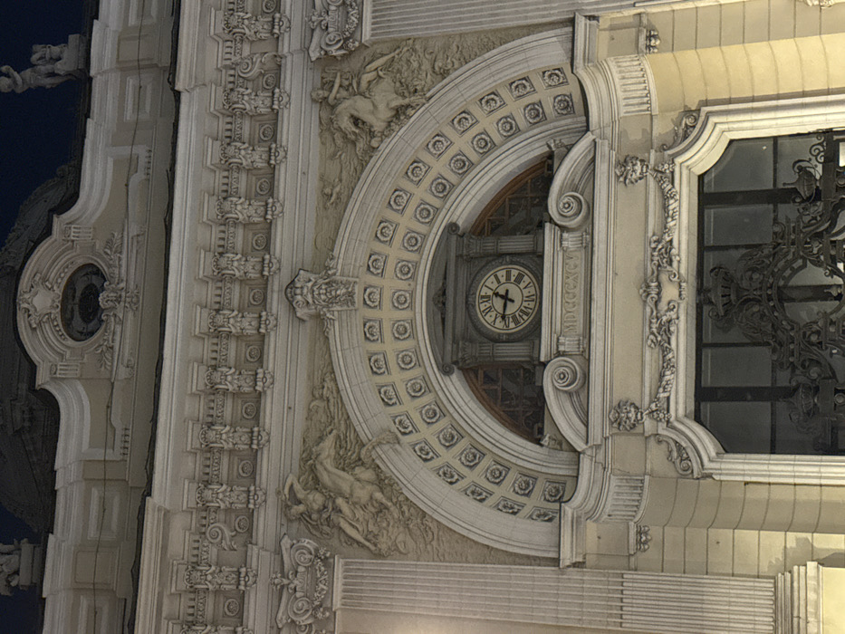The image displays a detailed, illuminated section of the ornate Neo-Baroque facade of the Petrovsky Passage building, located in Moscow, Russia, observed at night.
The central feature is a large arched recess, set within a light-colored, highly decorated wall. Within this arch, a circular clock with Roman numerals displays a time of approximately 10:15 or 10:20. Directly beneath the clock, the Roman numerals "MDCCCXCV" (1895) are inscribed. The arch itself is adorned with a repetitive pattern of square and circular decorative elements.
Flanking the arch are two large, stucco relief sculptures. The left relief depicts a figure (likely male, possibly a Triton or Centaur) riding a mythical marine creature or horse amidst swirling water. The right relief shows a male figure, possibly a warrior or classical deity, astride a rearing horse.
Above these reliefs and the arch, a prominent horizontal frieze runs across the facade, densely decorated with numerous small, stylized human figures or caryatids. Further architectural ornamentation, including cornices and additional sculptural details, extends upwards. A small, indistinct sculptural figure is partially visible against the dark night sky on the building's uppermost right edge within the frame.
Below the main arched section, a dark, ornate metal grille or gate covers a window or entrance. This gate features intricate scrollwork and a central crown motif. The entire facade is lit by artificial lights, casting shadows that enhance its three-dimensional decorative elements. No people or active interactions are visible.