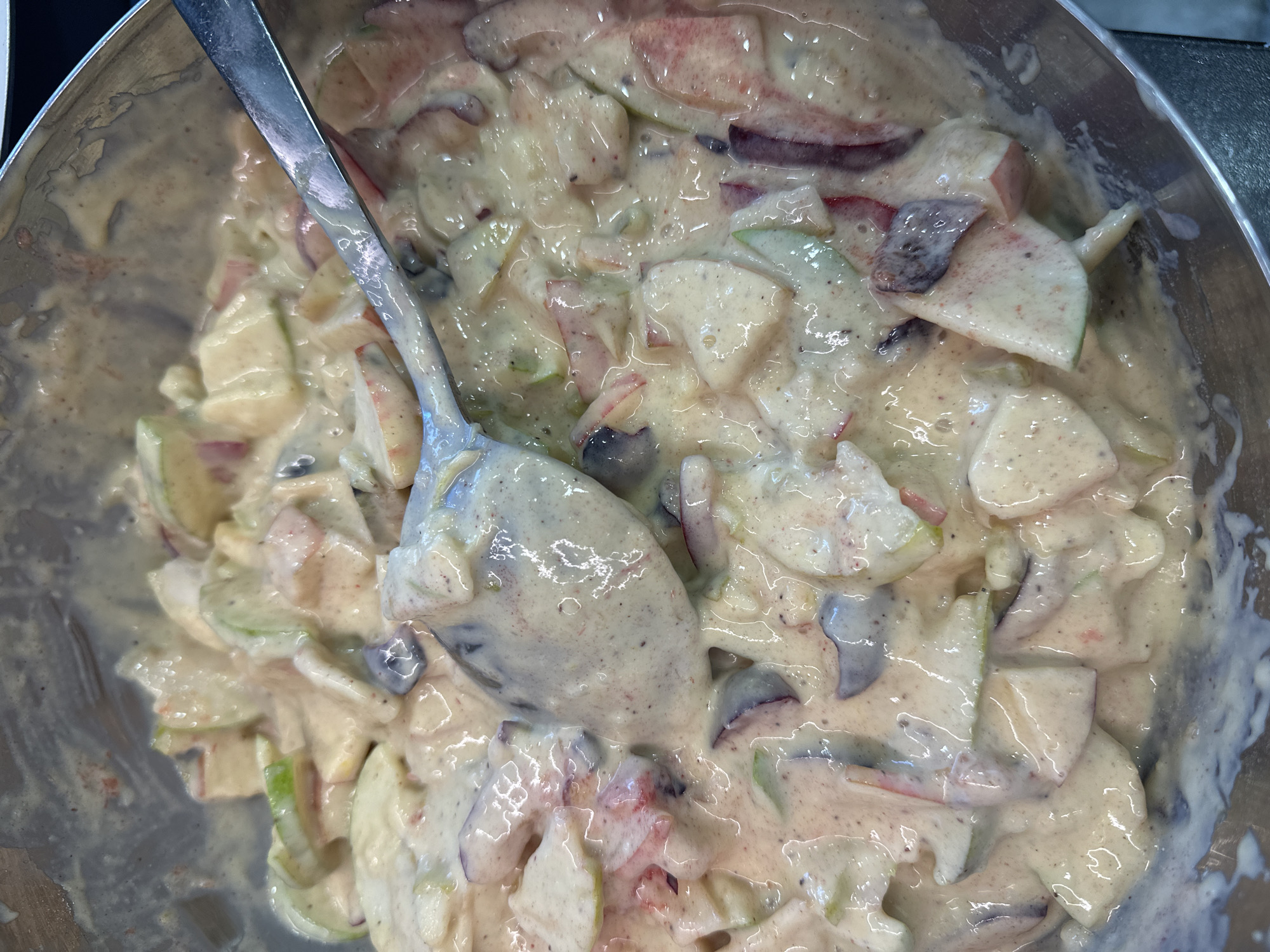 The image shows a close-up, overhead view of a metal bowl filled with chopped apples and a creamy dressing. A spoon is resting in the bowl, scooping up some of the mixture. The apples appear to be a mix of red and green varieties, cut into bite-sized pieces. The dressing is a pale yellow-white color with visible flecks of spices. The overall impression is of a prepared dish, likely a fruit salad. The image does not provide any information about its location in the city of Nevinnomyssk, Russia, or any contextual details.