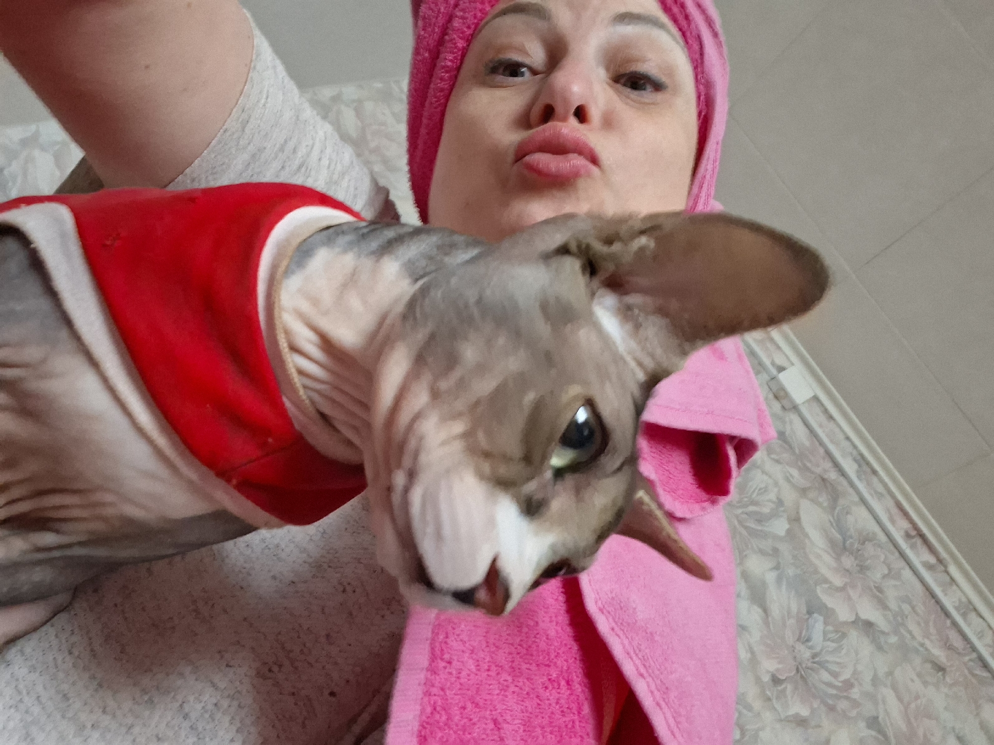 The image depicts a close-up, selfie-style shot of a person and a Sphynx cat. The person, on the left, is wearing a pink towel wrapped around their head and has their lips pursed. Their face is partially obscured by the cat. The cat, positioned centrally and to the right, is looking to the right and is wearing a red garment around its neck. The background appears to be an indoor setting with textured wallpaper. There are no discernible elements in the image that indicate a specific location in Забалоцкі сельскі Савет, Belarus.