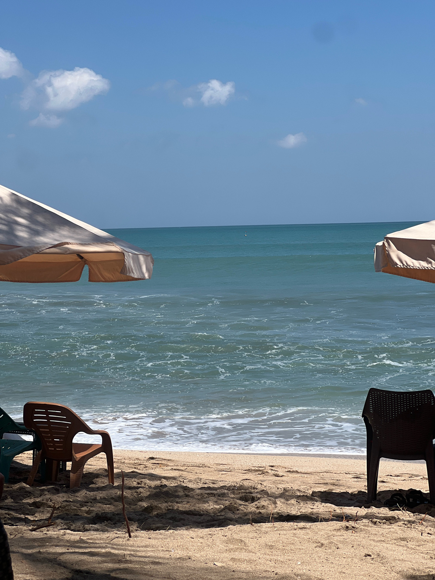 This is a medium shot of a beach scene in Kuta, Indonesia. Two beach umbrellas, one on the left and one on the right, are partially visible, casting shadows on the sand. Beneath the umbrellas, there are beach chairs. On the left, a brown plastic chair is positioned facing the ocean, with the back of a green chair visible to its left. On the right, a dark brown plastic chair is visible from the back. The sand is a light brown color, with small sticks and debris scattered on it. The ocean is a clear turquoise color with white waves breaking on the shore. The sky is a bright blue with a few scattered white clouds. The scene is peaceful and serene, conveying a sense of relaxation.