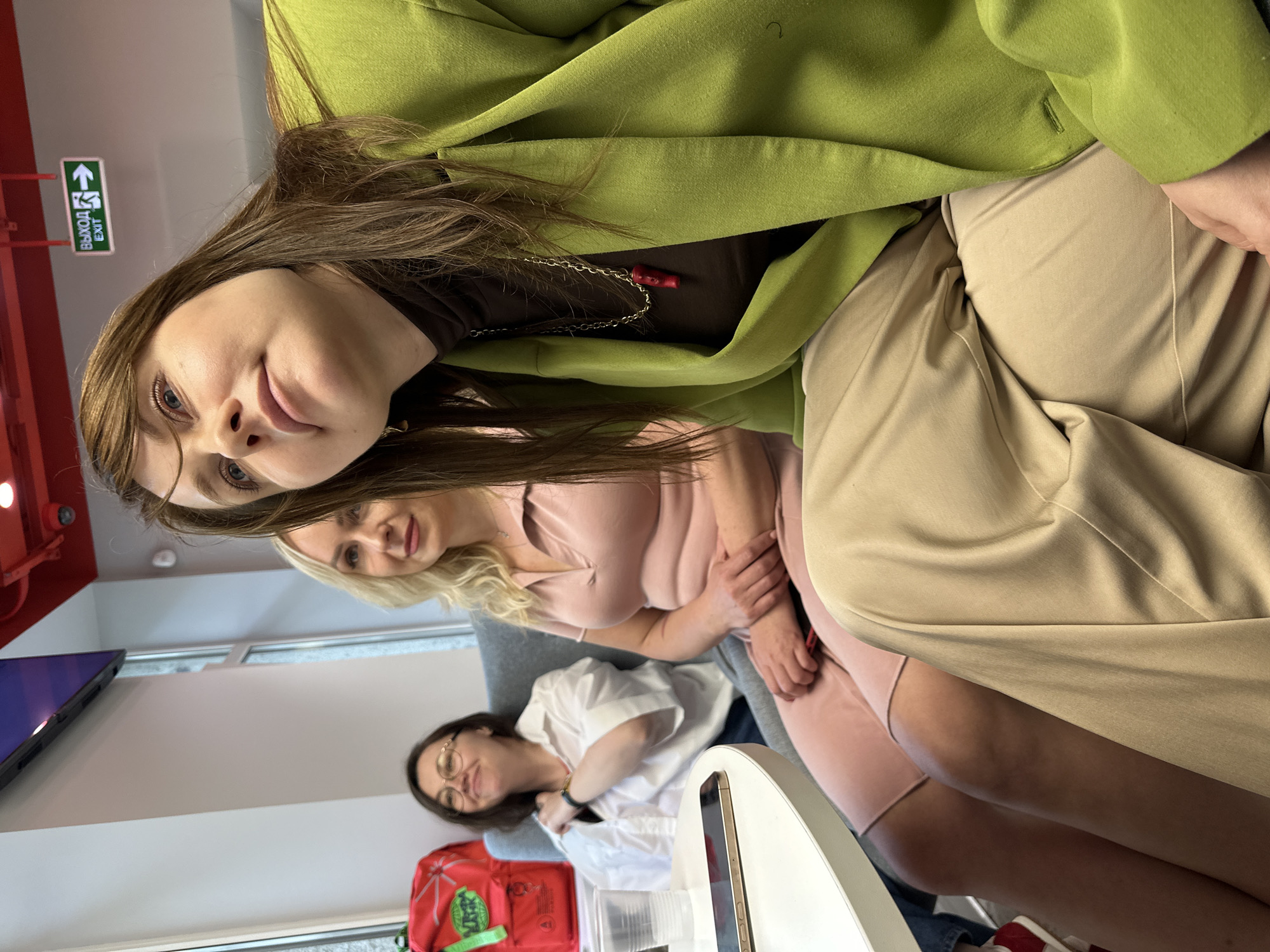 The image depicts three women seated on a sofa in what appears to be a modern, indoor space. The woman in the foreground, on the right, is wearing a lime green blazer and beige pants, and is looking upwards and to the right. She has a dark brown turtleneck underneath and a silver chain with a red pendant. The woman in the middle is wearing a pink dress and has blonde hair. The woman on the left is wearing glasses and a white shirt, and is smiling and looking to her left. A white table is in front of the woman on the left, with a phone and a plastic cup on it. Behind the woman on the left, a red backpack with green accents is visible. Above and behind the women, a sign with "ВЫХОД EXIT" and an arrow pointing to the right is affixed to a red ceiling structure. The overall environment suggests a commercial or public indoor area, potentially a cafe or a waiting area, possibly within a modern building in Sochi, Russia, given the Cyrillic text on the exit sign.