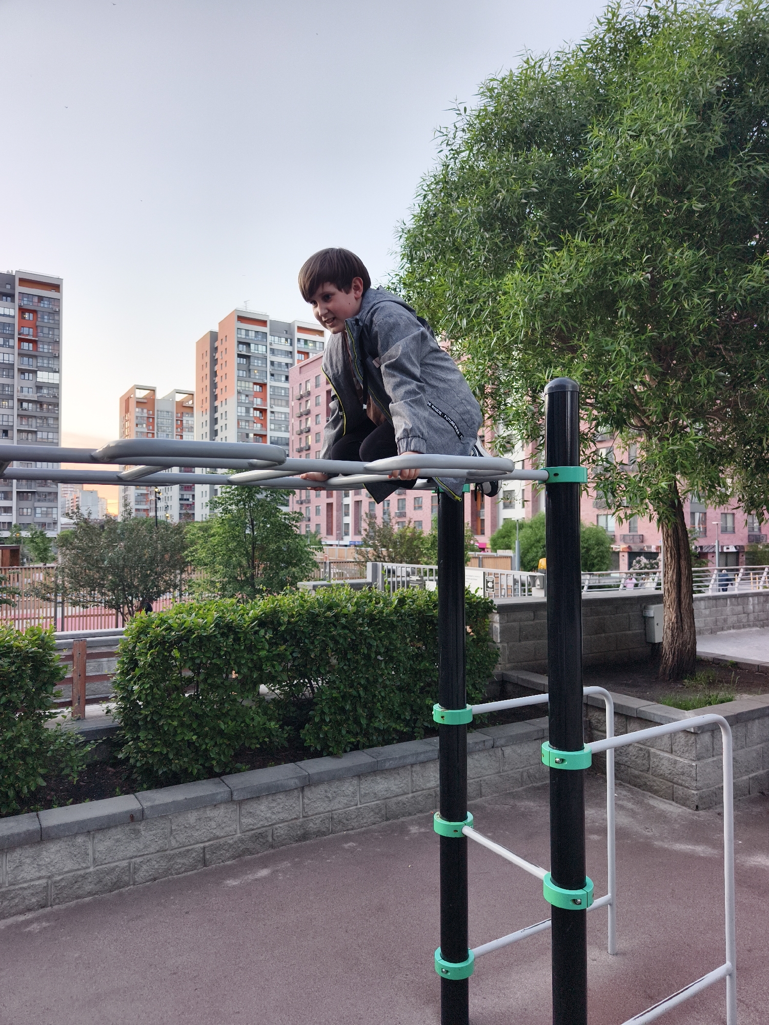 A young boy is captured in an active pose on an outdoor playground's horizontal ladder (monkey bars) in Tyumen, Russia. He has short brown hair and is wearing a gray denim jacket over dark clothing. His face beams with a wide, joyful grin, suggesting excitement and engagement in his play. On the cuff of his jacket sleeve, faint white text reads "WE INSIDE. FUTURE...".
The boy is balanced on one of the gray metal horizontal bars, gripping it with his hands. The playground equipment features sturdy black vertical poles secured by bright light-green circular clamps, which hold the gray bars in place. The ground directly beneath the equipment is a reddish-brown surface, likely asphalt or a rubberized play surface. To the right, part of another white metal exercise structure is visible.
In the midground, a low grey stone wall is topped with a well-maintained green hedge. Behind the hedge, several trees with green foliage are present, including a large tree with narrow, flowing leaves on the right side of the image. The background is dominated by modern, multi-story residential buildings, varying in color from gray to orange and pastel pink, typical of a developed urban environment. The sky above is clear and bright but pale, indicating either early morning or late afternoon light, which casts a soft, even illumination over the entire scene. The image conveys a cheerful moment of childhood play against a contemporary city backdrop.