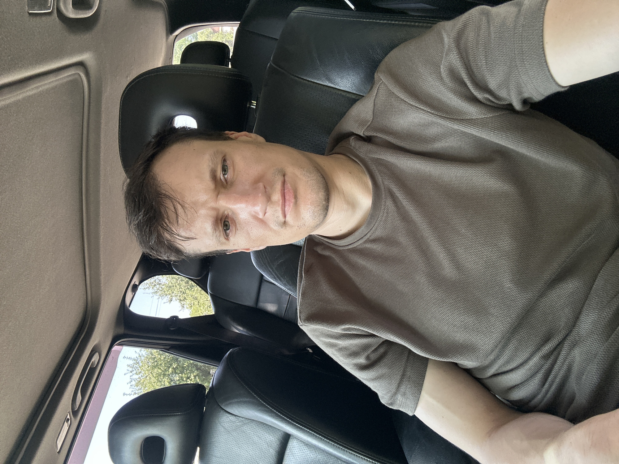The image is a selfie taken from inside a car, with the man's face and upper body filling the frame. He is seated in the driver's seat, with black leather seats visible on either side. The interior of the car appears to be a SUV, with a sunroof overhead. Outside the car, trees and a clear sky are visible through the windows. The man is wearing a brown t-shirt and has short brown hair. He has a neutral expression on his face. No specific location in Makiv, Ukraine can be identified from the image alone.
