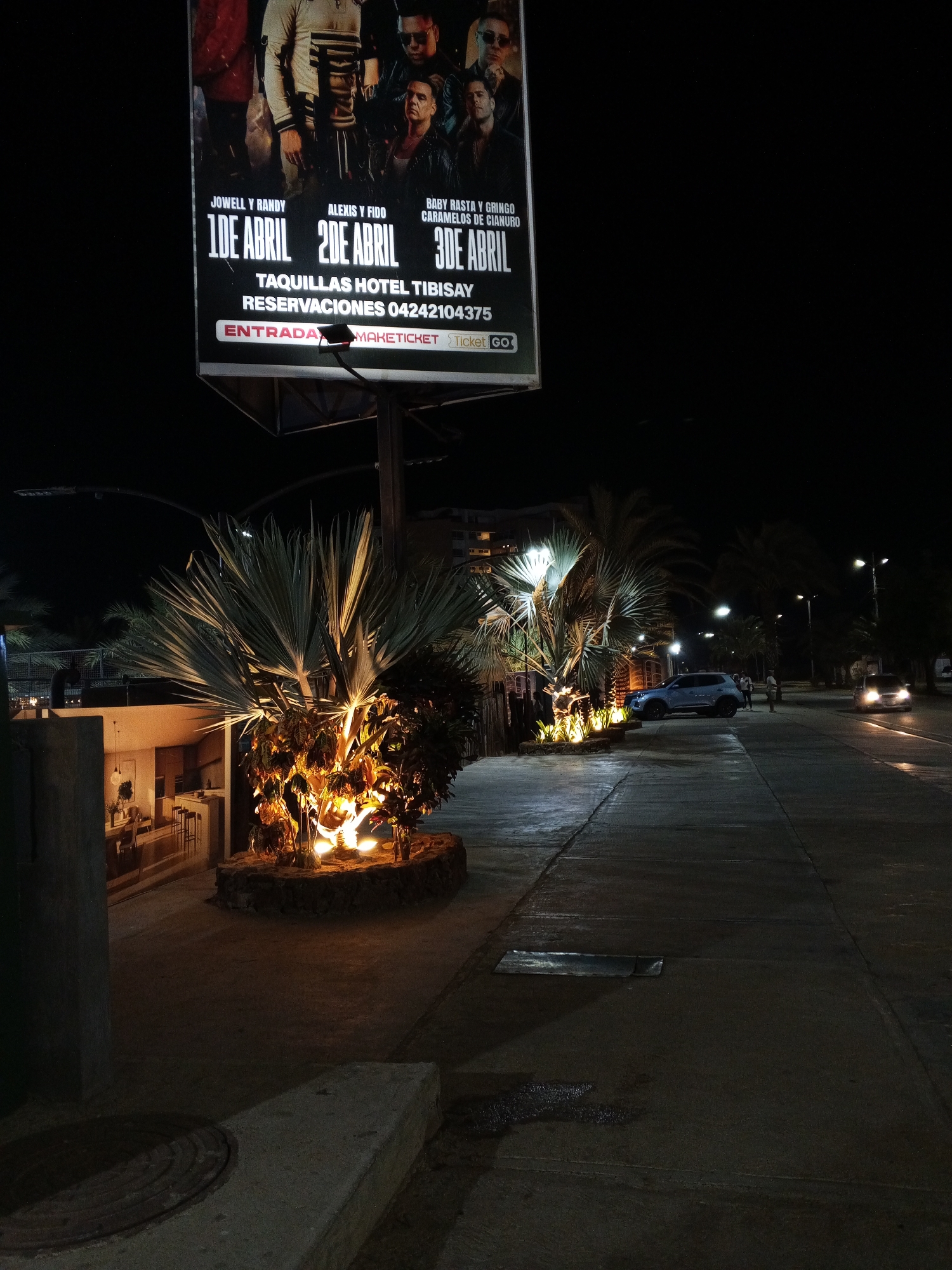 The image depicts a nighttime scene in Pampatar, Venezuela. A large advertising billboard for a musical concert, featuring the names "Jowell y Randy," "Alexis y Fido," and "Baby Rasta y Gringo Caramelos de Cianuro" with dates of "1DE ABRIL" "2DE ABRIL" and "3DE ABRIL," is prominently displayed. The text "TAQUILLAS HOTEL TIBISAY" and "RESERVACIONES 04242104375" is visible under the dates, along with "ENTRADA" and "MAKETICKET Ticket GO" logos.
Below the billboard, illuminated trees and shrubbery line a sidewalk, and some nearby structures are partly visible. Cars are seen on the road. The scene is mostly dark, with the light sources being street lights, car headlights, and the illuminated signage and trees. The sidewalk appears wet in some areas.