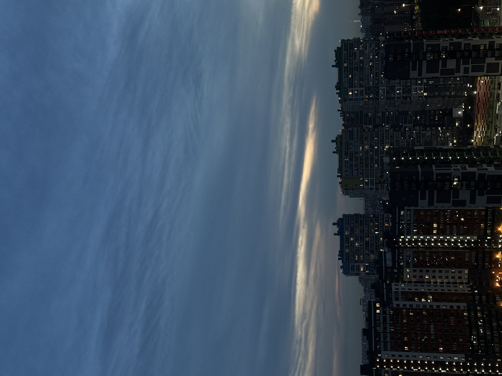 This is a nighttime aerial photograph of a densely populated residential area in Feklistovo, Russia. The image captures a cluster of modern, multi-story apartment buildings, illuminated by the warm glow of interior lights and streetlamps. The sky above is a deep twilight blue, streaked with wispy clouds that catch the last vestiges of daylight. The architecture of the buildings is varied, with some featuring darker facades and geometric patterns, while others are lighter in color and have more uniform window arrangements. The sheer density of the buildings suggests a large urban development. No people are visible in the frame. The overall impression is one of a bustling, contemporary city at dusk.