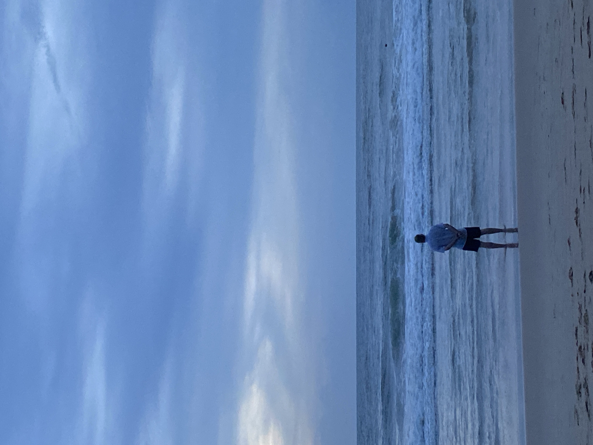 A man is standing in the surf on a beach. The sky is overcast and blue. The location is Choeng Thale, Thailand.