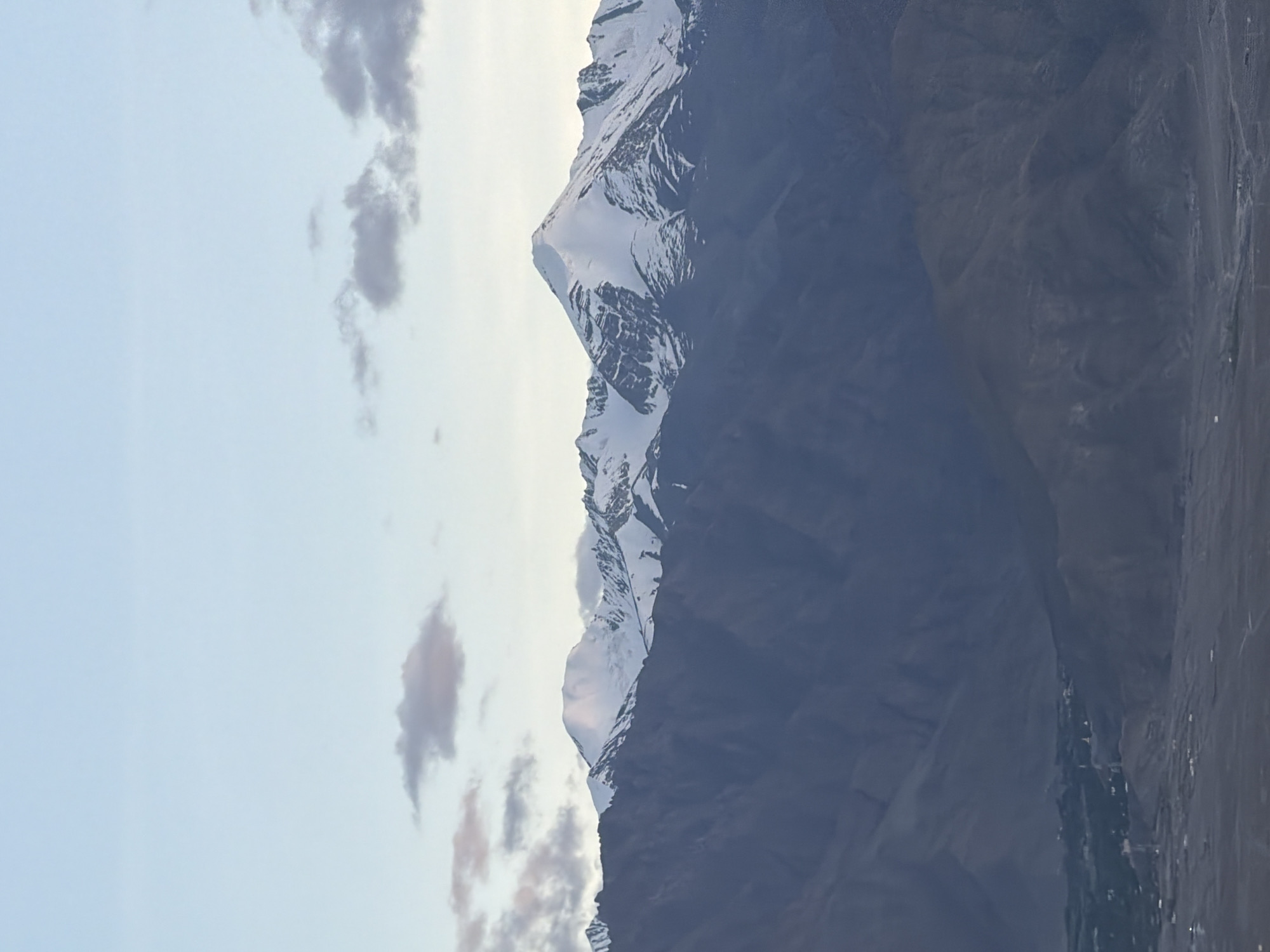 The image shows a panoramic view of the majestic Himalayan mountains surrounding the city of Leh, India, under a pale blue sky with scattered clouds. The foreground is dominated by the rugged, dark brown slopes of the lower mountains, gradually receding into the distance. Further back, the imposing peaks of the higher Himalayas rise, their summits dramatically capped with pristine white snow. The contrast between the barren, arid lower slopes and the snow-covered peaks is striking, showcasing the diverse geography of the region. In the lower part of the frame, a small settlement or village is visible, nestled in the valley amidst the foothills, hinting at human habitation in this vast and awe-inspiring landscape. The overall impression is one of immense natural beauty, solitude, and the grandeur of the high-altitude environment of Leh.