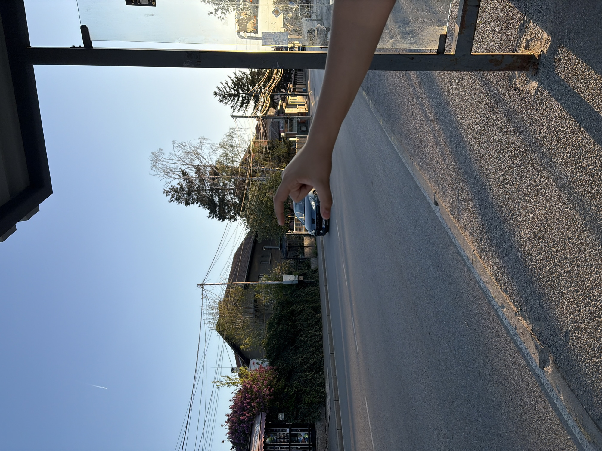 The image depicts a sunny street scene in Sofia, Bulgaria, captured from inside a bus stop shelter. The perspective is rotated 90 degrees clockwise from its natural orientation, showing the sky on the left and the ground on the right. When viewed upright, a human arm and hand extend from the upper right of the frame, performing a playful optical illusion by appearing to 'pinch' or 'grasp' a distant, dark-colored car on the road.
The scene features a two-lane asphalt road with a curb and a rough-textured sidewalk to its right. Long shadows stretch across the ground and parts of the bus stop structure, indicating late afternoon or early evening during the golden hour. The bus stop shelter itself has a dark metal frame and clear glass panels, with some unreadable text visible on a sticker.
In the background, typical residential buildings or houses with tiled roofs are visible, interspersed with various trees – some deciduous with bare branches and some evergreen pines. Overgrown bushes and a patch of vibrant purple/pink flowering shrubs add color to the scene. Numerous utility poles and tangled power lines crisscross the sky and stretch along the street. A small, black vending machine stands near the flowering shrubs.
A dark car, possibly blue or black, is partially visible on the road, with its license plate "CA 059..." confirming the location as Sofia, Bulgaria. A white airplane contrail streaks across the clear blue sky. A sign with the word "TAXI" is also discernable on a pole in the mid-ground. The overall impression is one of a casual, everyday moment in an urban or suburban setting, with a touch of lightheartedness due to the hand gesture.
