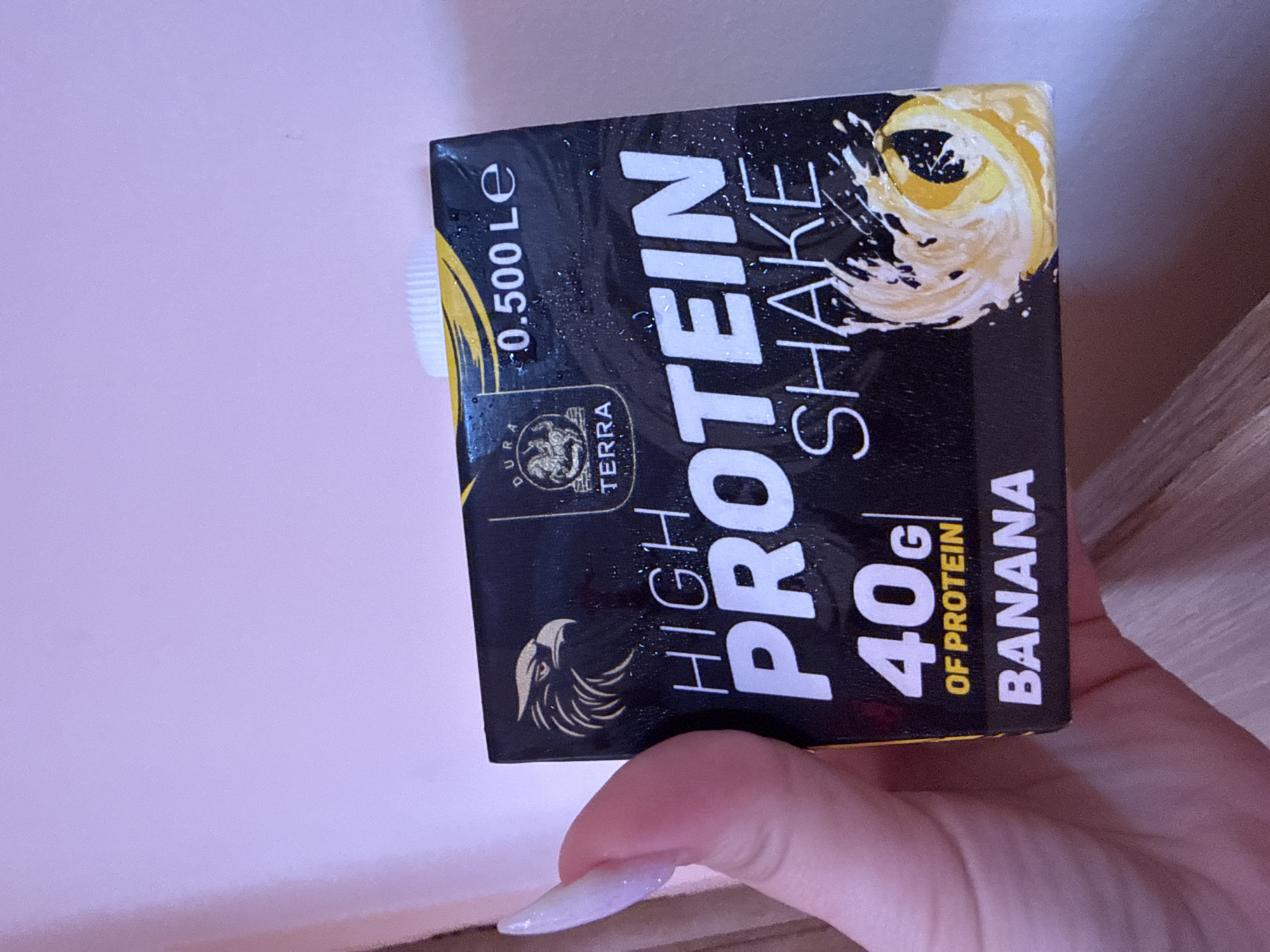 The image displays a person's hand holding a black protein shake carton against a light purplish-pink background, with a portion of a wooden surface visible on the right. The carton is oriented horizontally due to the image's rotation.
The primary object is a black protein shake carton, its surface covered with small water droplets, suggesting it is cold. Large white text on the carton reads "HIGH PROTEIN SHAKE". Below this, "40g OF PROTEIN" is stated, and the flavor is clearly marked as "BANANA" in yellow letters. The carton's volume is indicated as "0.500 Le" at the top left corner. A brand logo featuring an eagle or griffin is visible alongside the partially visible text "DUA TERRA", and another stylized eagle head logo is present lower on the carton. The top right corner of the carton features a graphic of swirling yellow and white liquid, likely representing the banana flavor or the shake itself. A white plastic screw cap seals the carton.
A hand with long, pale, possibly iridescent or light pink polished fingernails is seen holding the carton from beneath. The scene appears to be indoors, under artificial lighting. The activity is simply the act of holding the protein shake. The background is mostly a plain, pastel-colored wall or surface. No specific time of day or emotional cues are apparent.