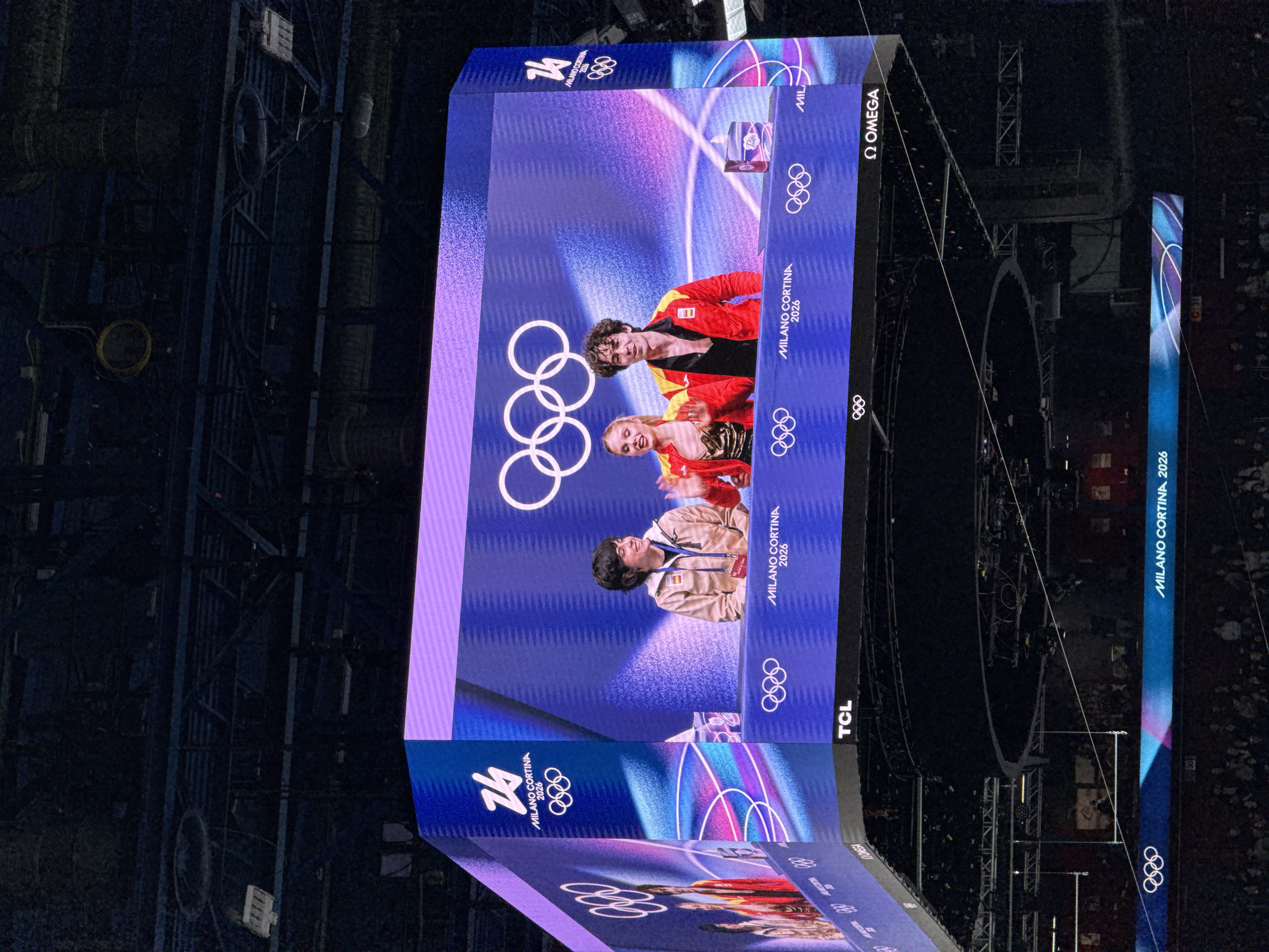 The image shows a large LED screen in a dark indoor venue, possibly an arena. The screen displays images and logos related to the 2026 Milano Cortina Winter Olympics, including the Olympic rings. It features the logo on each section of the screen. People in athletic wear, possibly athletes, are pictured in the center. The screen also displays brand logos such as Omega and TCL. In the background, there is stage rigging.