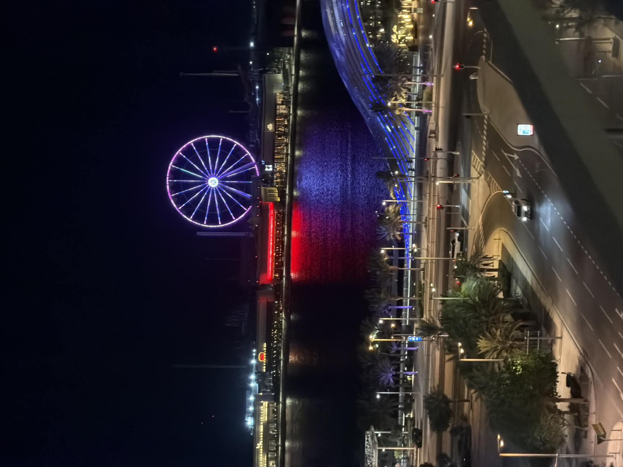 This elevated night photograph depicts a bustling urban waterfront at Bluewaters Island, Dubai, UAE. The prominent Ain Dubai Ferris wheel dominates the upper center, illuminated in vivid purple, pink, and light blue radial patterns, casting reflections on the dark, rippled water below. Along the opposite bank, a series of brightly lit commercial buildings line the promenade, one clearly displaying "The Cheesecake Factory" sign, while another structure on the right projects a strong red light onto the water.
In the mid-ground, a multi-lane asphalt road with clear lane markings curves from left to right, bordered by numerous tall palm trees and modern streetlights that cast a warm glow. Several vehicles, including white and dark-colored cars, are present on the road, with headlights visible on moving vehicles and some stopped at red traffic signals. Small, indistinct figures of pedestrians can be seen on the sidewalks. A distinctive, modern architectural structure with multiple blue light strips arches over or alongside the water on the right side of the frame. The sky is a deep, clear black, contributing to the dramatic contrast with the vibrant artificial lighting throughout the scene.
