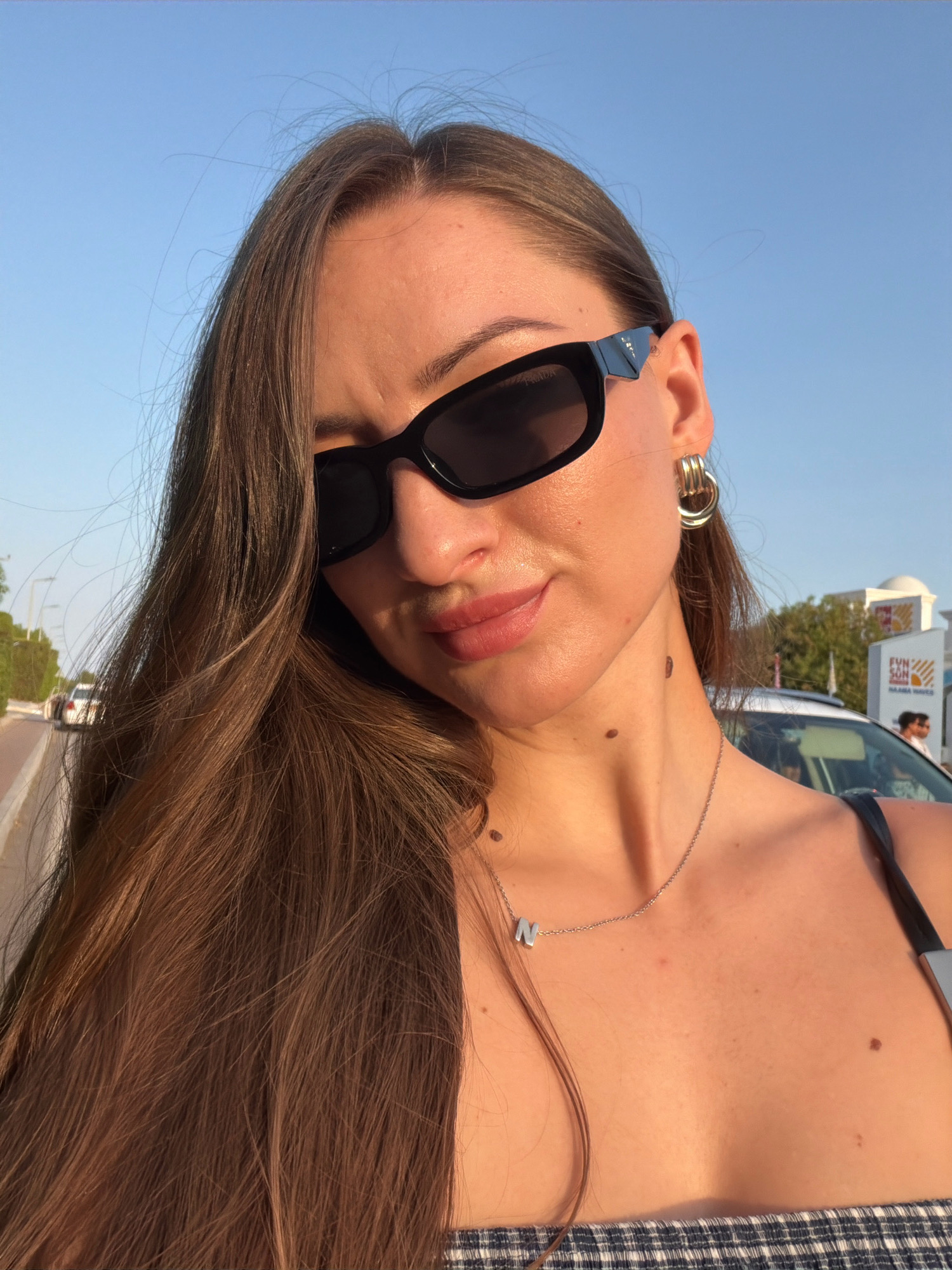 A woman with long brown hair and sunglasses poses for a selfie outdoors. She is wearing a plaid top and a silver necklace with the initial "N". In the background, there is a street with trees, a car, and a sign that reads "FUN SUN NAAMA WAVES". The scene appears to be in Sharm El Sheikh, Egypt, likely in the Naama Bay area, known for its tourist attractions. The lighting suggests it is late afternoon or early evening.