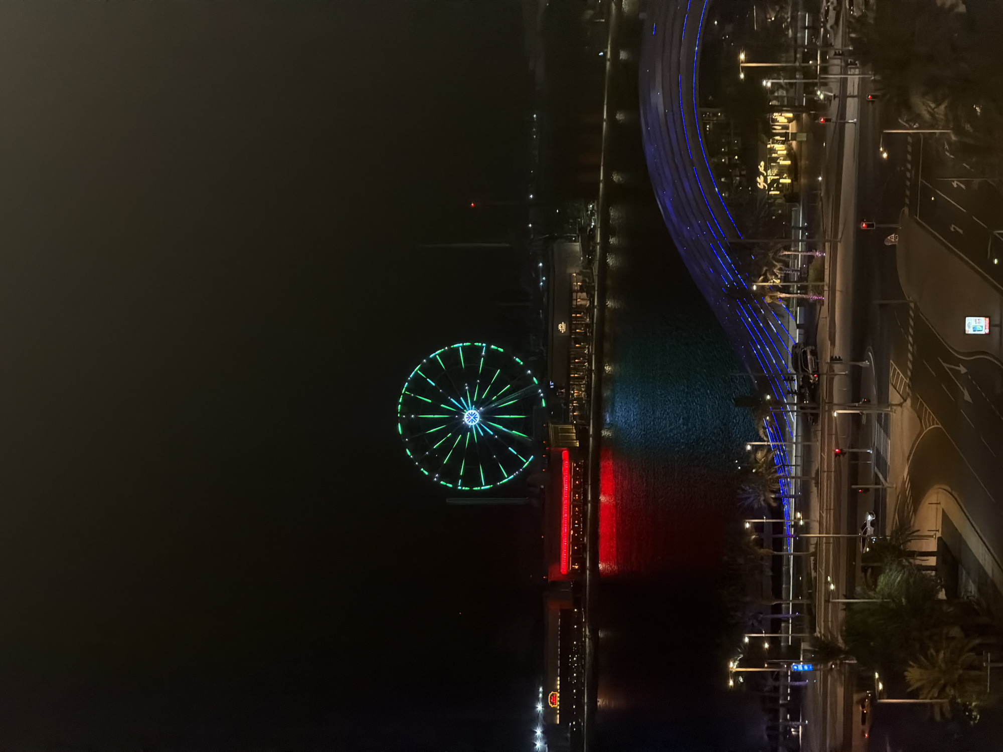 An aerial night view captures a waterfront city scene. A large Ferris wheel illuminated with green and blue lights dominates the background, positioned above a row of buildings. Red lights are reflected in the water, creating a striking contrast with the cooler tones of the Ferris wheel and the overall dark sky. In the foreground, a modern, arched bridge lit with blue lights spans across the scene, with palm trees and streetlights lining the roads below. Cars are visible on the road network, and the overall ambiance suggests a vibrant urban environment at night. The location is identified as Unknown, Unknown.