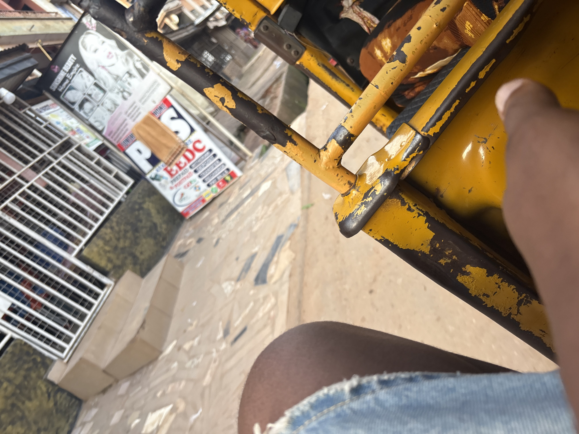 This image was taken in Onitsha, Nigeria, from the perspective of someone sitting in a yellow motorized tricycle (keke napep). The tricycle's weathered yellow paint, with areas of rust and black showing through, is prominent in the foreground. To the left, a person's leg clad in distressed denim shorts is visible. In the background, a building with a metal security grate over windows is seen, along with a tiled walkway. A poster advertising "Sunshine Beauty Home Empire" is affixed to the side of the building, displaying images of women's faces. The overall impression is an urban street scene in Nigeria.