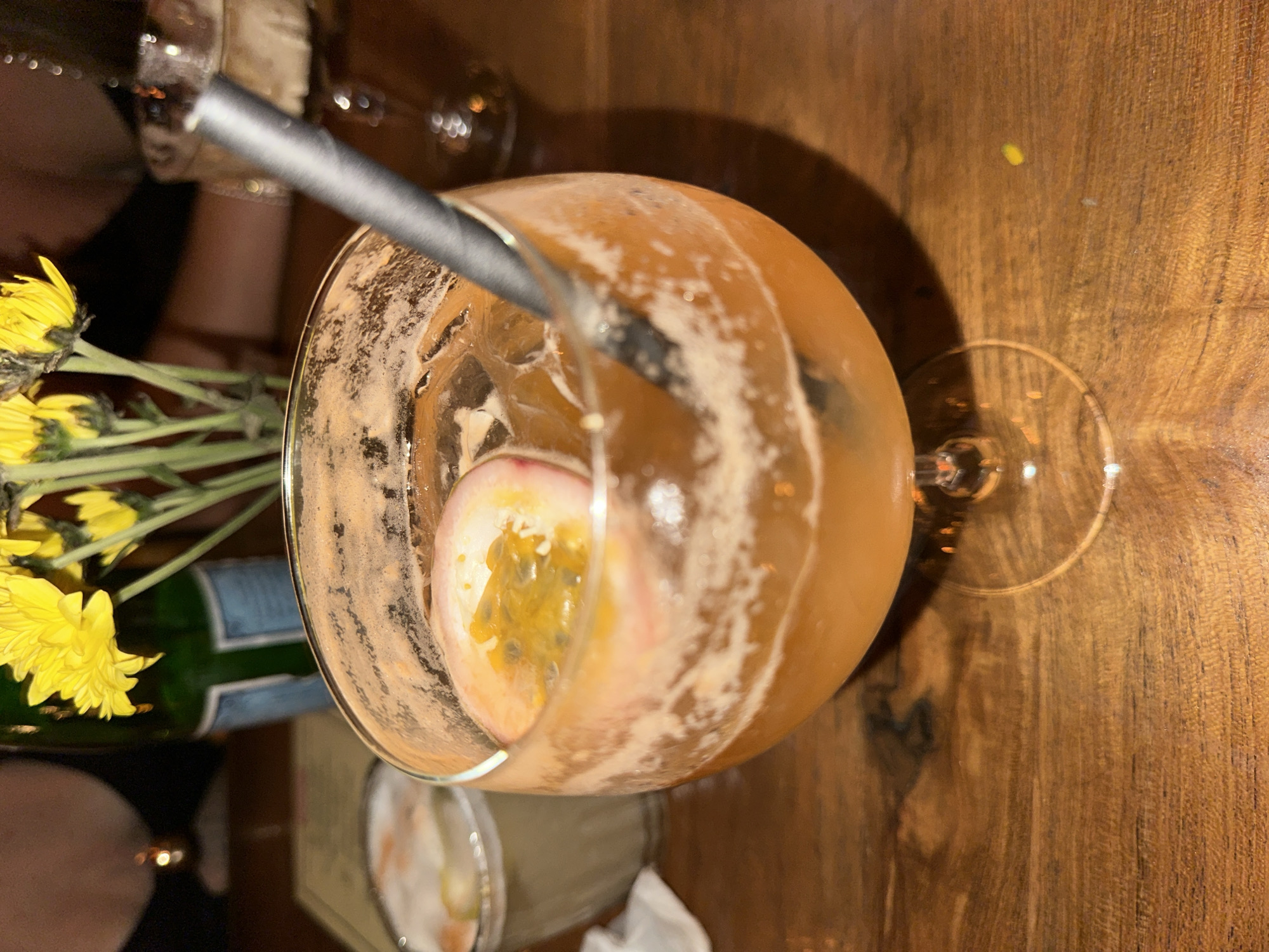 The image depicts a cocktail in a stemmed glass on a wooden table. The cocktail contains a passion fruit half and a straw. Adjacent to the glass, there is a vase with yellow flowers and a green bottle. The location is implied to be in Kuta Selatan, Indonesia. People are partially visible in the background.