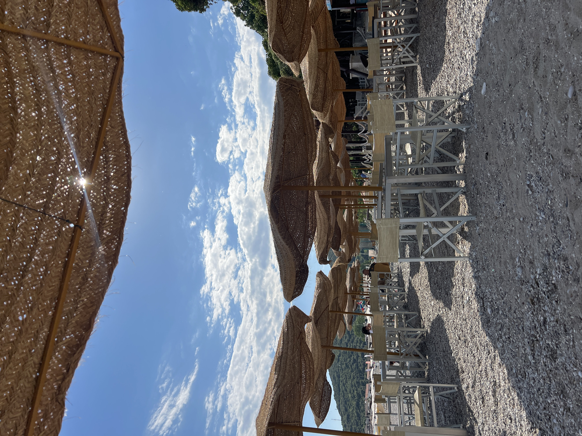The image shows a beach scene, likely in Prijevor, Montenegro, given the location request. Rows of beach umbrellas with straw-like canopies are visible, interspersed with white chairs and tables. The ground is covered with pebbles or coarse sand. A bright blue sky with scattered clouds provides a backdrop. Trees and some buildings are visible in the distance. The sun shines through the umbrella canopy in the foreground, causing a bright glare. People are present in the scene, seated in the chairs in the background.