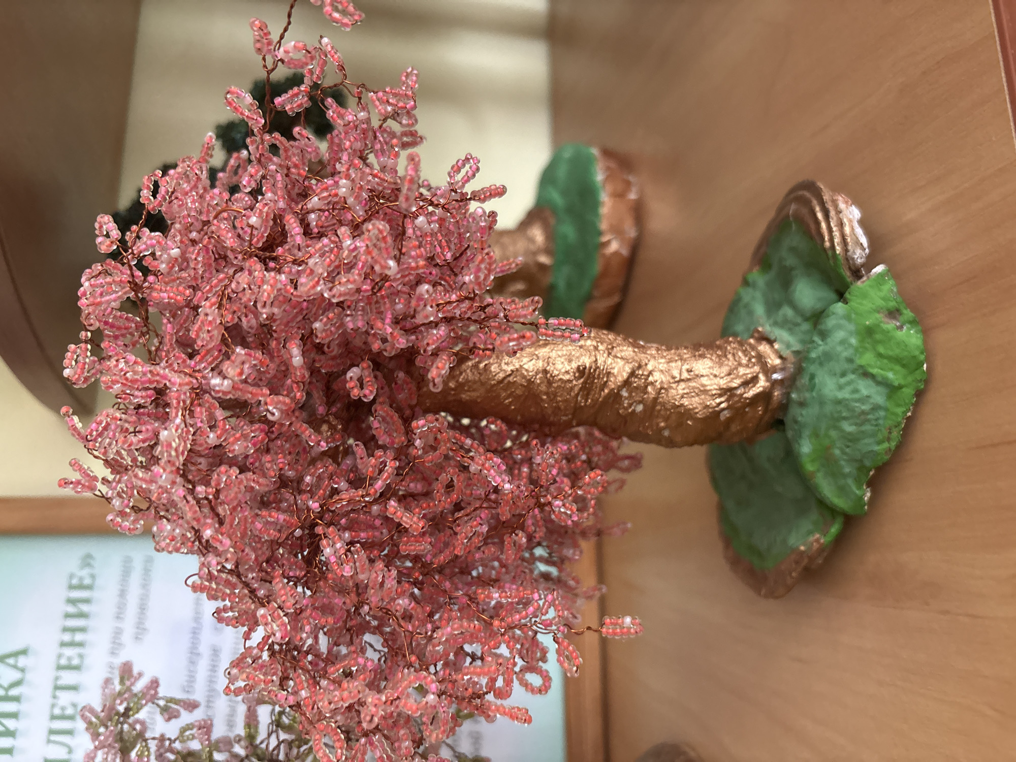 The image depicts a close-up, eye-level view of a small, handmade decorative tree made of pink and clear beads, with a gold-painted trunk and a green, irregularly shaped base. The tree is the primary subject and is in sharp focus. In the blurred background, a piece of paper with text in Cyrillic is visible, suggesting an indoor setting, possibly an educational institution or a craft exhibition. The text on the paper is partially obscured but appears to be a title or a descriptive passage. The overall impression is of a craft item displayed in a simple environment. The location of Ветрынскі сельскі Савет, Belarus is not directly discernible from the image itself, but the presence of Cyrillic text hints at a Slavic-speaking region, consistent with Belarus.