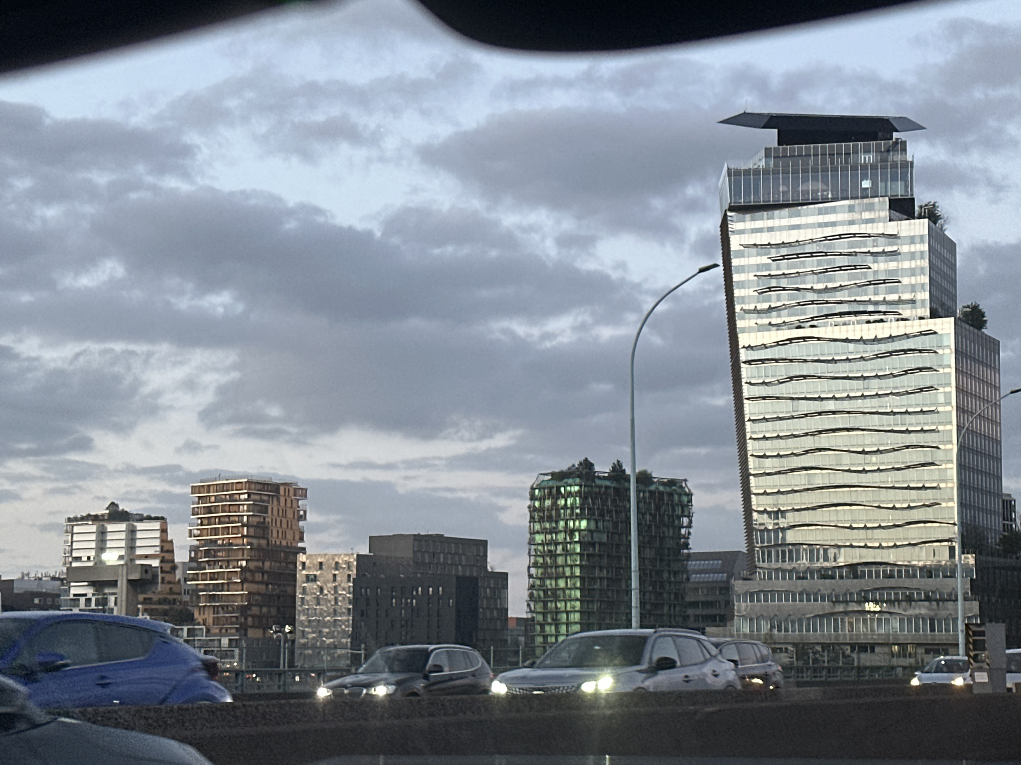 The image shows a cityscape with several modern buildings under a cloudy sky. The most prominent building is a tall, glassy skyscraper with a distinctive flat top. The building's facade has horizontal, wavy lines that give it a unique appearance. In the foreground, there are several cars on a road, suggesting that the photo was taken from a moving vehicle. Based on the architectural style of the buildings, the location is likely in the La Défense district of Paris, France. There are no visible people in the image.