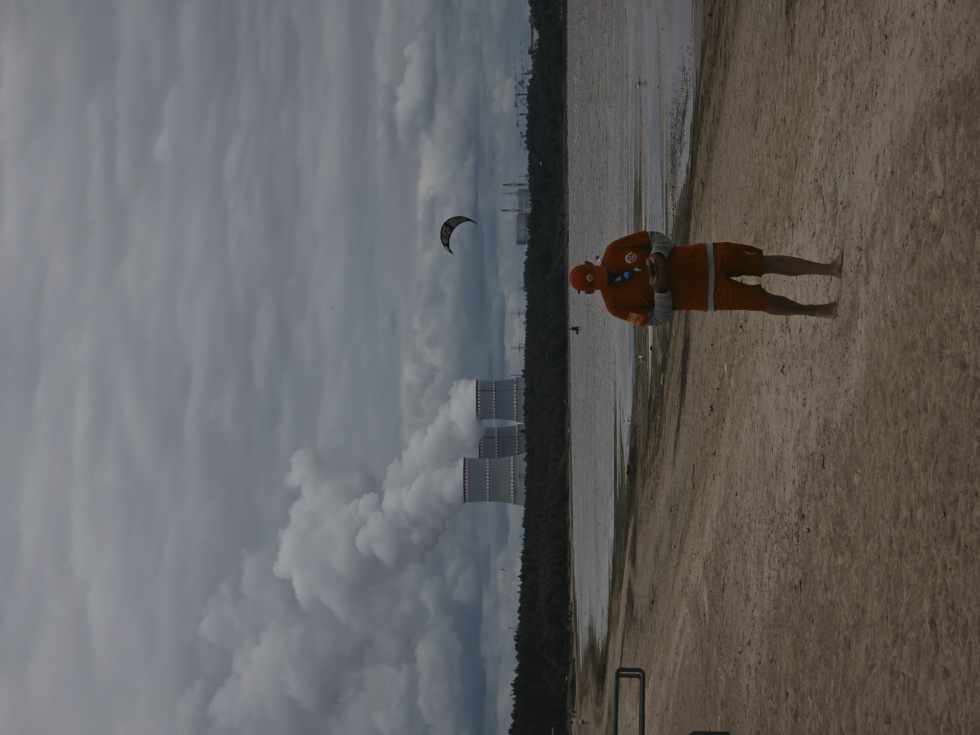 A person wearing an orange shirt and shorts stands on a sandy beach. The person is holding a phone in their hands and appears to be looking down at it. Behind the person, a body of water stretches out towards a line of trees. In the distance, several large, industrial cooling towers are visible, emitting plumes of white smoke into a cloudy sky. A kite is also visible in the sky, in the shape of a crescent moon. The weather appears overcast. The image is oriented vertically, with the beach and the person on the right side of the frame, and the water, trees, and cooling towers on the left. The sandy beach is textured with small pebbles and debris. The water appears still, reflecting the cloudy sky. The cooling towers are massive structures, suggesting an industrial or power plant location. The overall mood of the image is somewhat somber due to the overcast sky and the industrial elements in the background. The text "ROK" is visible on the person's shirt.