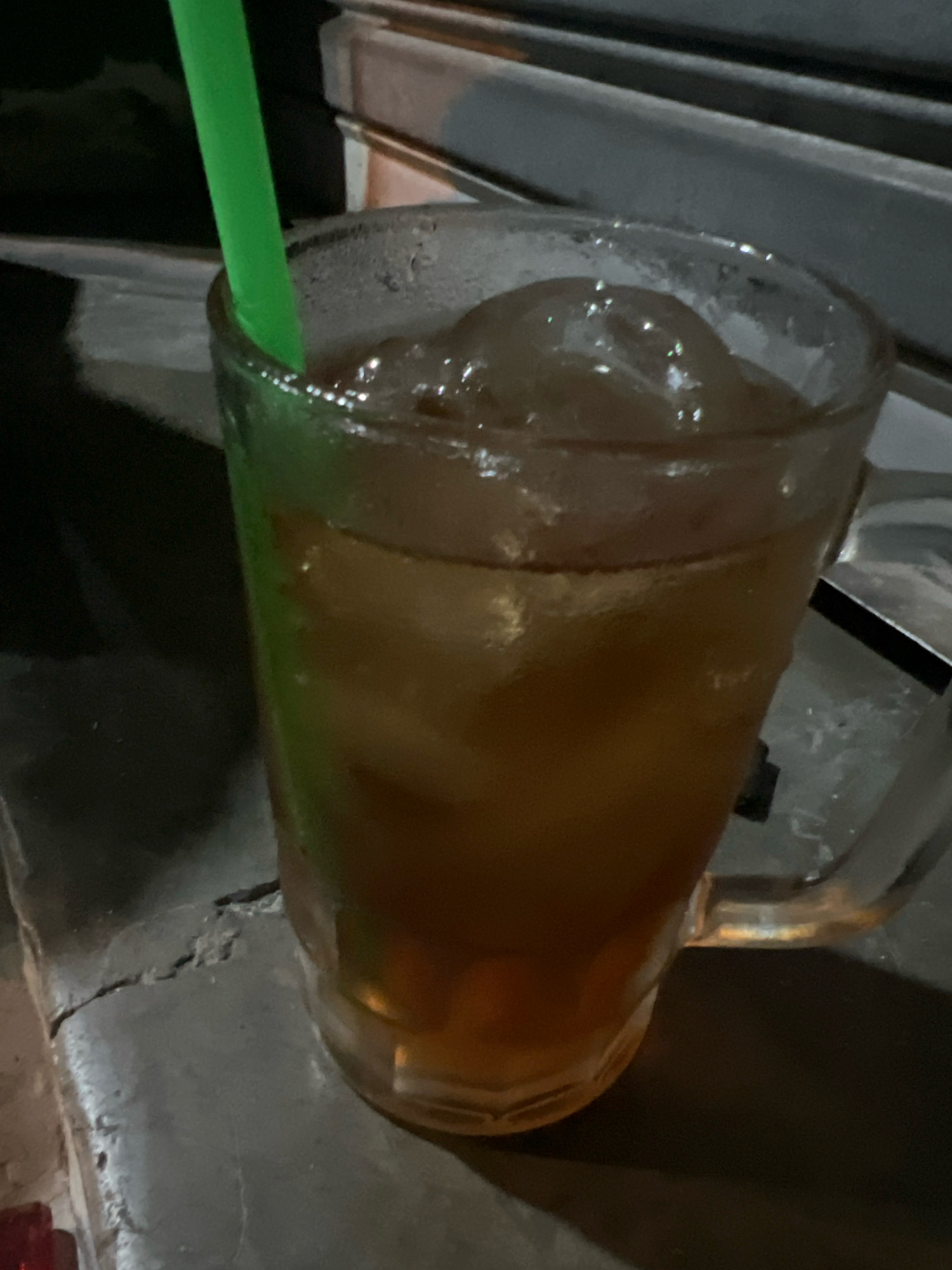 A close-up, eye-level shot shows a tall, clear glass mug filled with iced tea and a green straw. The mug, which has a handle, is positioned on a rough, gray surface that appears to be concrete with cracks. The iced tea is a deep amber color with numerous ice cubes visible. Condensation is visible on the outside of the glass. In the background, a dark, out-of-focus area suggests an outdoor setting, possibly a street or roadside. The lighting is dim, indicating it might be evening or nighttime. This scene is likely located in Tlogoadi, Indonesia, given the context provided.