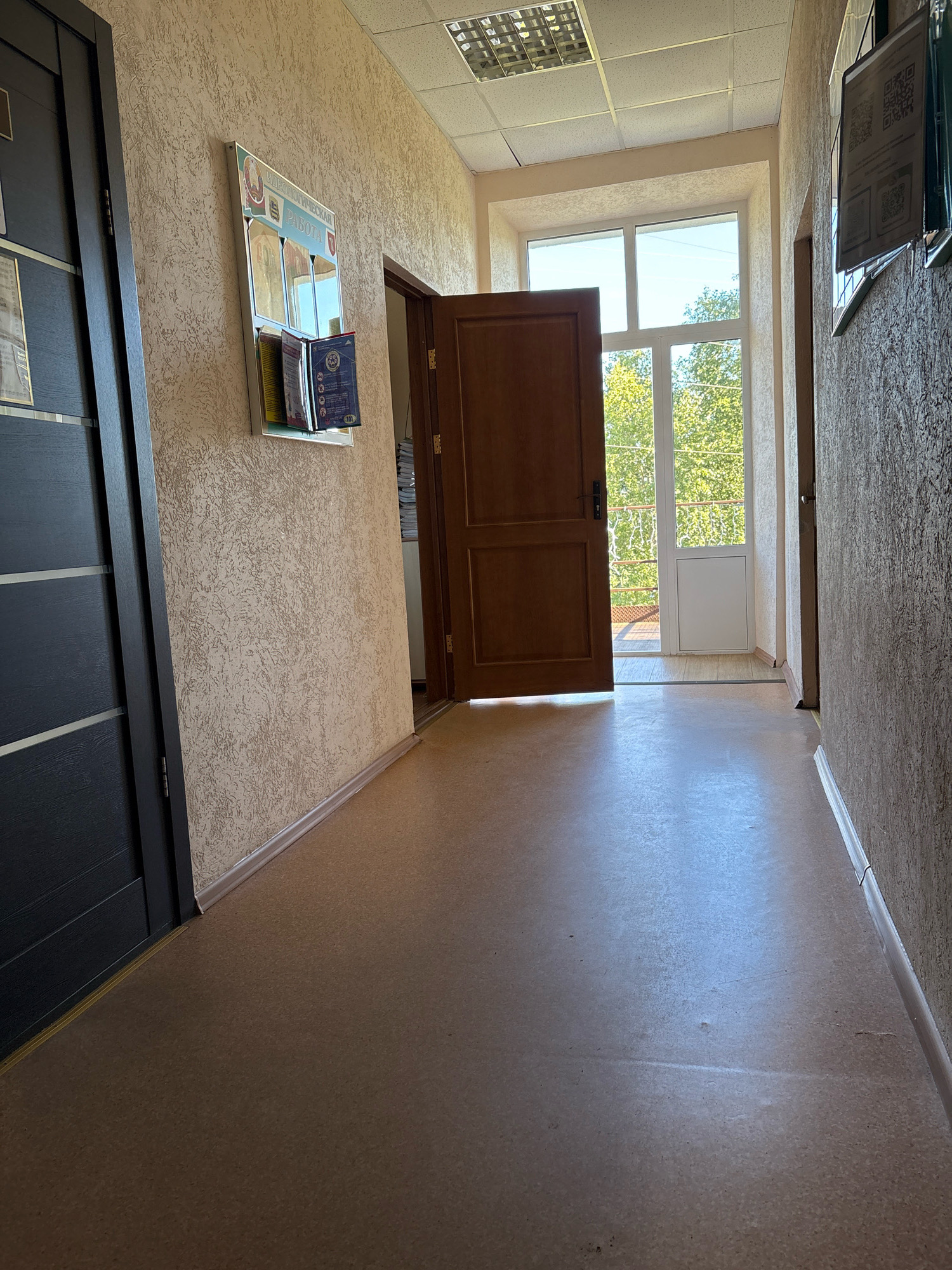 The image shows a hallway with a cork floor and textured beige walls. On the left wall, a dark grey door is visible, with a bulletin board attached to the wall next to it. The bulletin board displays several papers and brochures. Further down the hallway, a wooden door is open, revealing a view of trees and sky through a window and a balcony with a railing. Another door is visible on the right wall. The ceiling has fluorescent lighting. The scene appears to be an interior common area, possibly in an office building or residential complex.