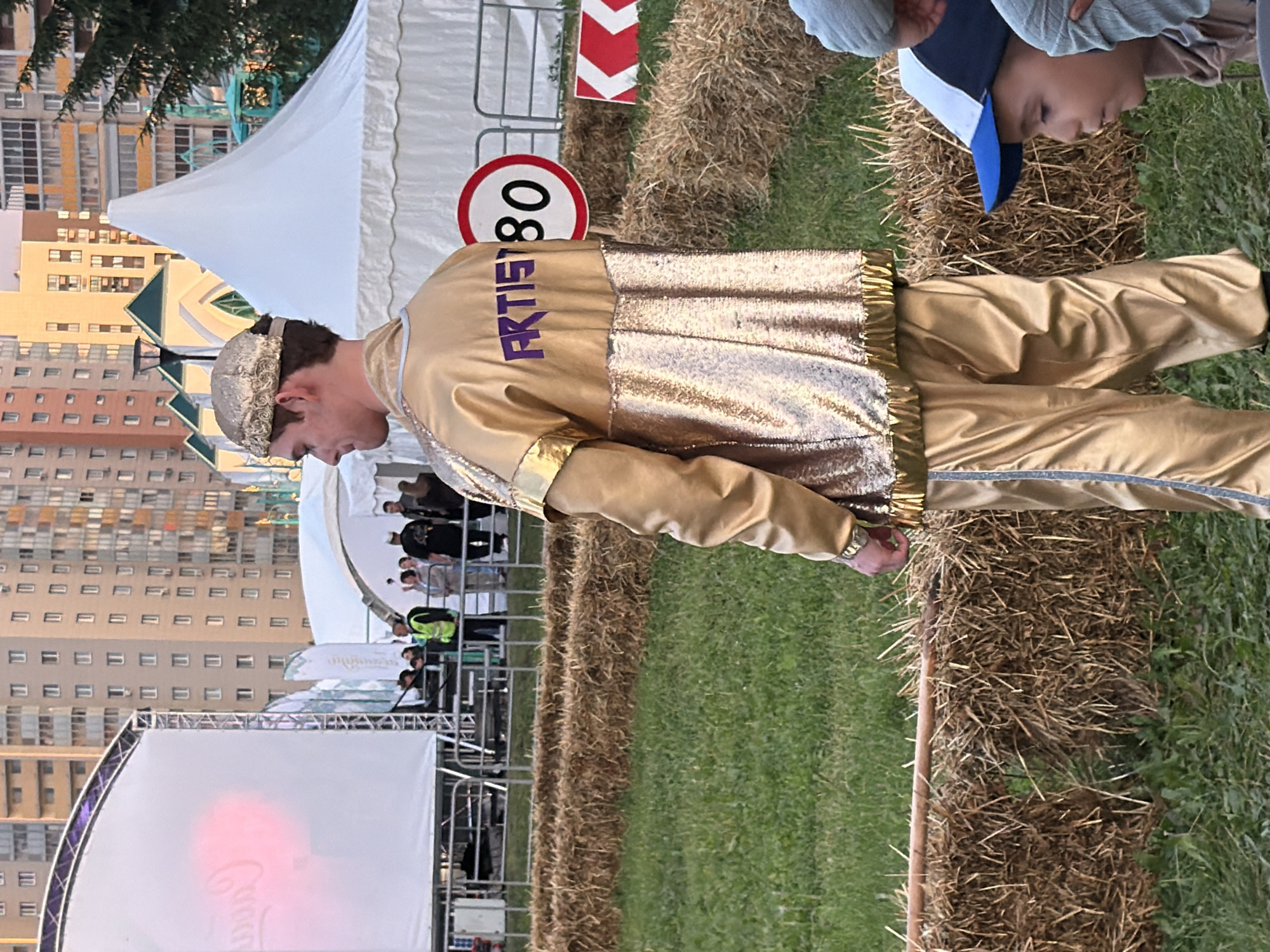 In an outdoor urban setting, a young man in a gold sequined tracksuit and a gold cap stands facing away from the camera. The tracksuit has the word "ARTIST" in purple lettering on the back. He is standing in front of a row of hay bales and a grassy area. Behind him, a speed limit sign indicating "80" is visible, along with a white tent structure and tall apartment buildings in the background. A large white screen with a pink projection is also present on the left side of the frame. Several people are milling around in the background, and two children are partially visible in the lower right corner of the image.