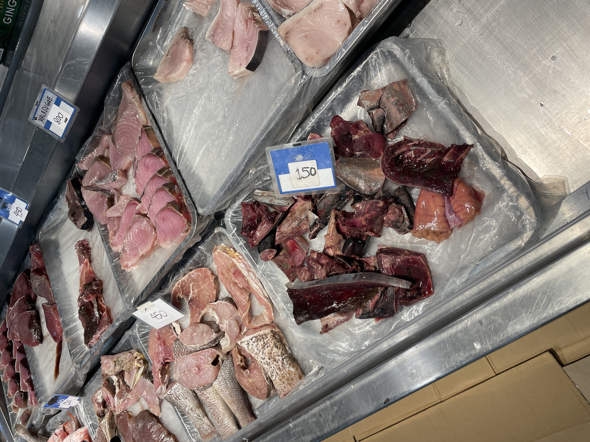 The image shows a display of raw fish at a market in Davao City, Philippines. The fish is laid out on trays filled with ice and plastic wrap. Various types of fish are visible, including steaks of pinkish-white fish, whole fish, and cut pieces of dark red fish. Some of the fish have price tags attached, with prices like "450" and "800" visible. The market appears to be indoors, with stainless steel countertops and shelving. The overall impression is that of a bustling marketplace.