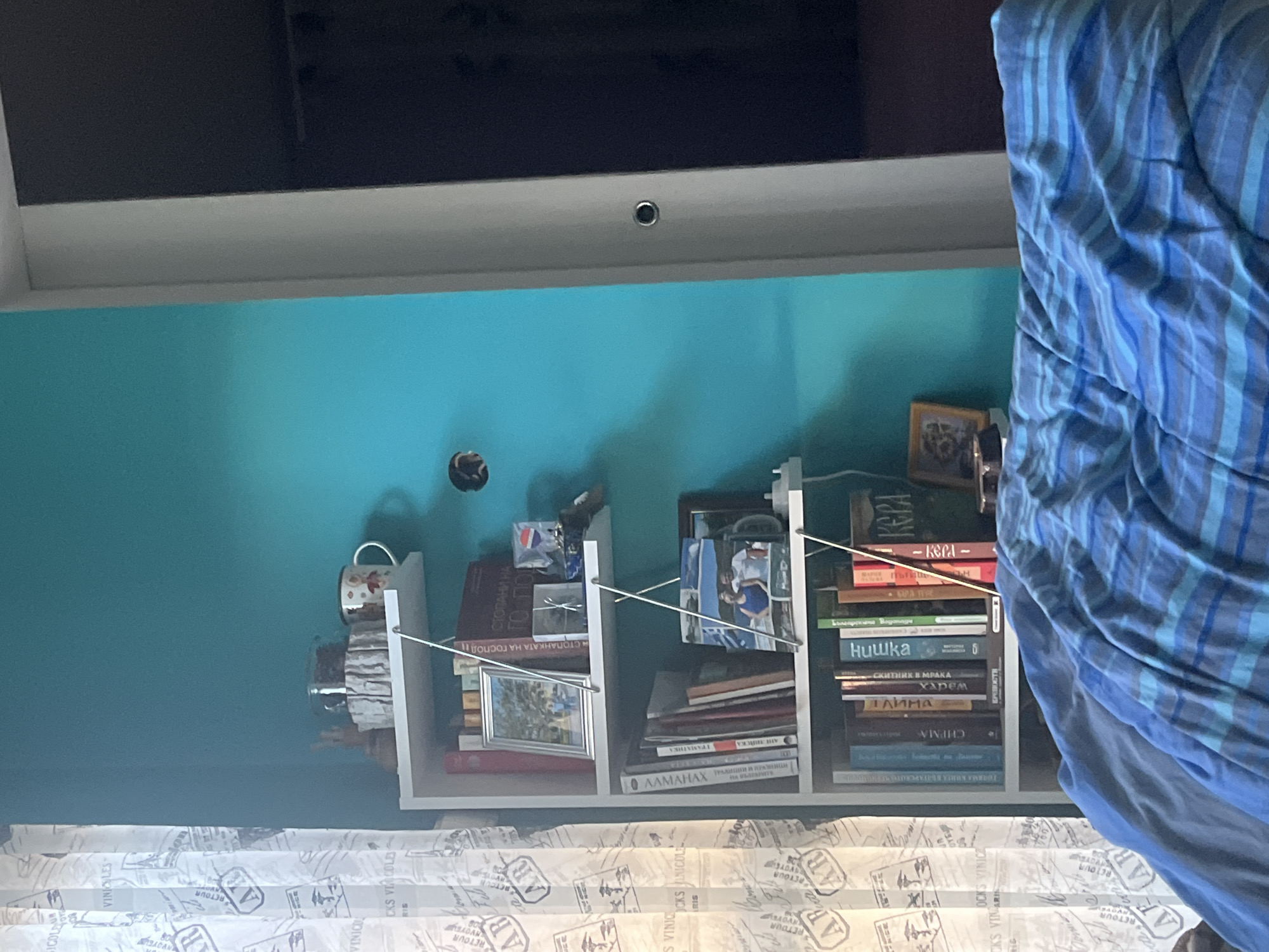 The image shows a room with a teal wall. On the wall is a white shelf filled with books and various objects, including cups and framed pictures. A blue striped blanket partially obscures the right side of the shelf. Above the wall is a dark cabinet. A white and beige patterned curtain is visible along the bottom edge of the image. The OCR text includes Bulgarian text "СТОПАНКАТА НА ГОСПОДН" which translates to "THE FARMER'S WIFE". Additionally "АЛМАНАХ ТРАДИЦИИ И ПРАВНИЦИ" meaning "ALMANAC OF TRADITIONS AND PRACTICES". The location is likely an indoor setting. There is no evidence to indicate that the image was taken in Pokrovnik, Bulgaria.