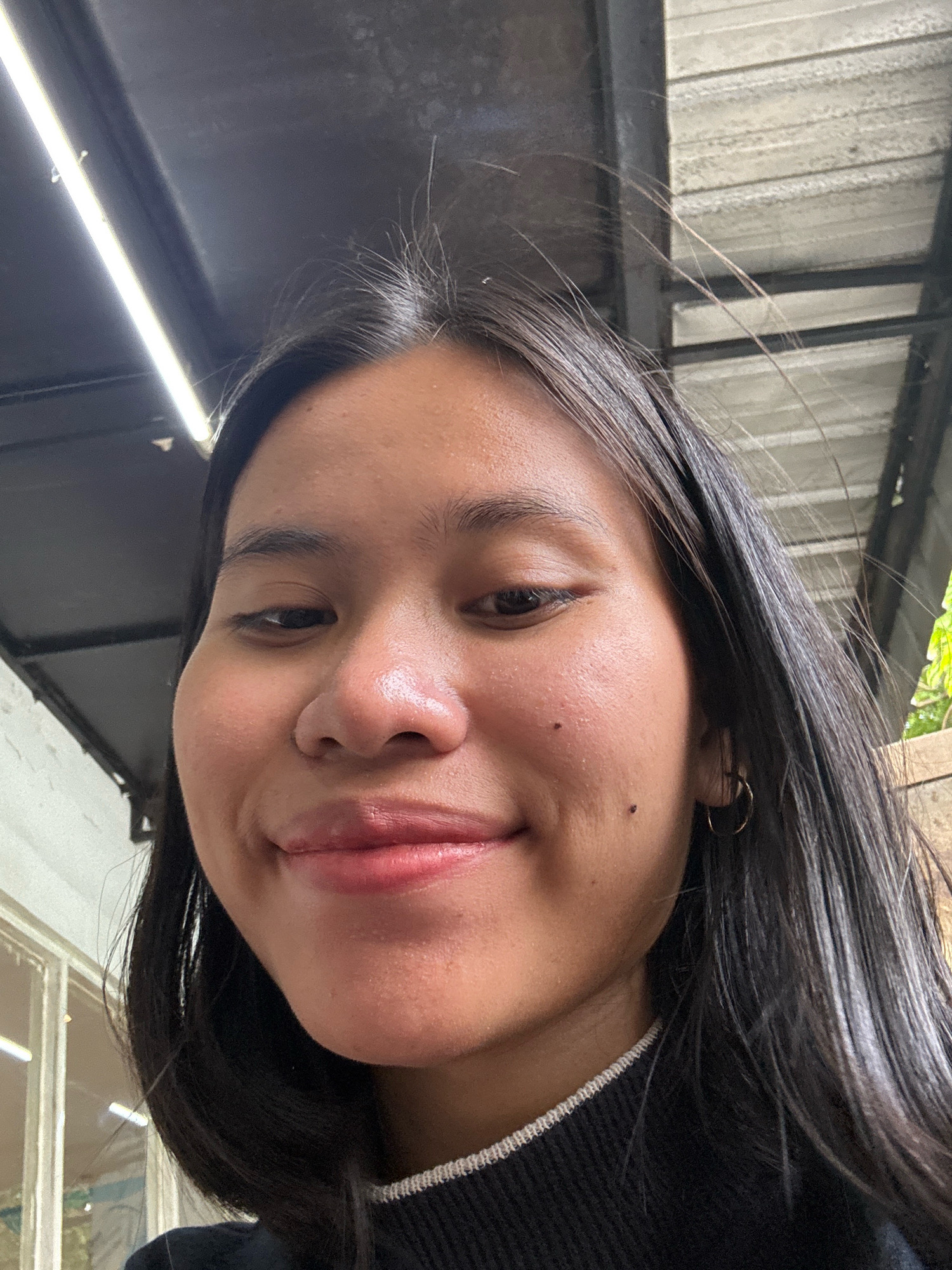 A close-up, slightly upward-angled selfie of a young East Asian woman with long, dark hair. She is wearing a black sweater with a white trim at the neckline. She has a subtle smile, revealing a portion of her upper teeth. Two small dark moles are visible on her right cheek. A thin, gold hoop earring is present in her right ear. The background suggests an outdoor or semi-outdoor setting, possibly a covered walkway or patio, with a corrugated metal roof and artificial lighting visible. The image was taken in East Jakarta, Indonesia.