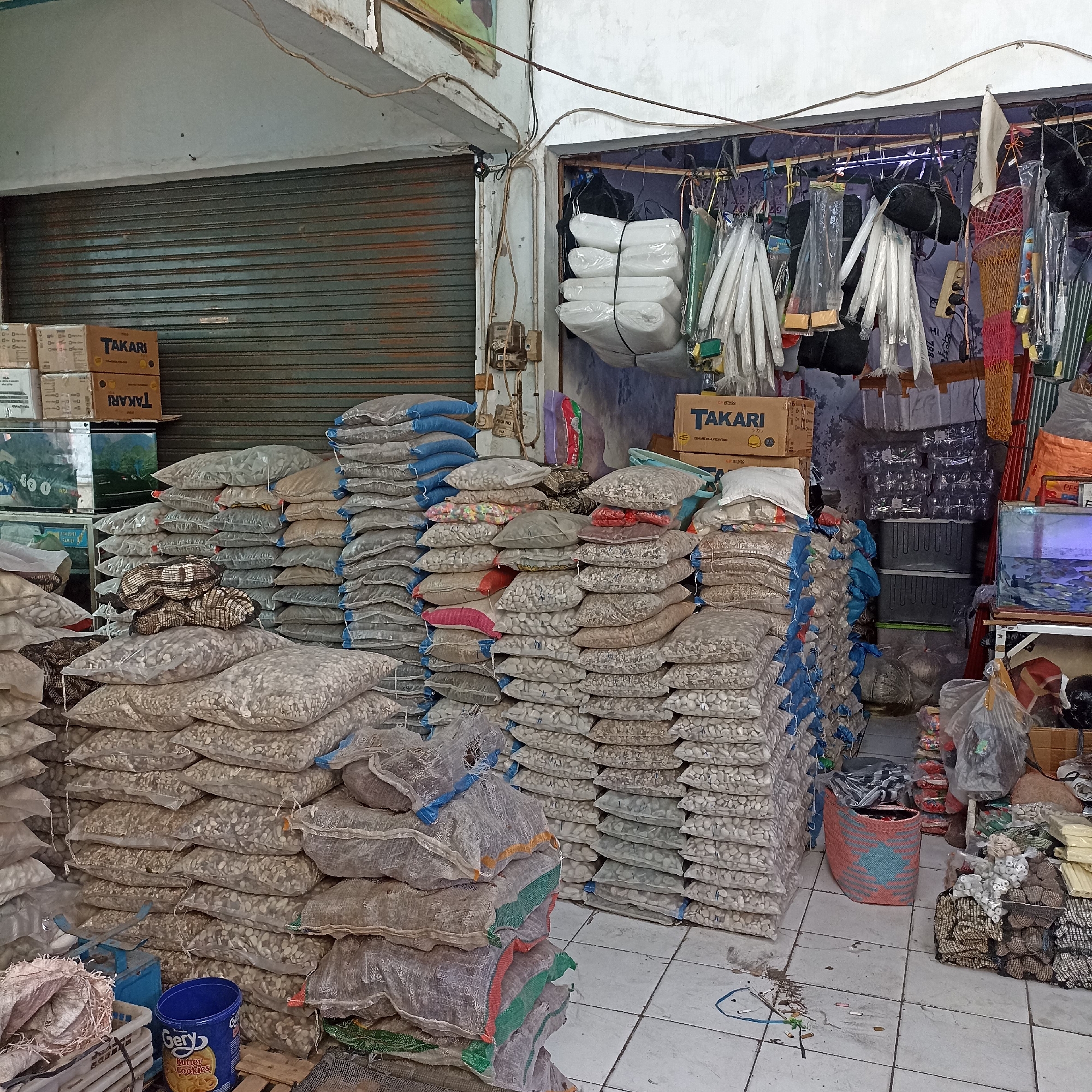 The image depicts a market stall in Magelang, Indonesia, likely selling pet supplies or aquarium accessories. In the foreground, large stacks of clear plastic bags filled with small, light-colored pebbles are prominent. Some bags have colored netting or tie-offs. A blue bucket labeled "Gery Butter Cookies" is visible near the bottom left. Behind these stacks, more bags of pebbles continue to fill the space.
To the left, two glass aquariums are partially visible, one containing water and fish. Several cardboard boxes with the "TAKARI" logo are stacked on shelves above the aquariums, suggesting they might contain fish food or related products.
The background reveals more shelving and hanging items. Several white, cylindrical objects wrapped in plastic and some dark, bundled items are suspended from a rack. A roll-up metal door, partially closed and appearing weathered, dominates the left side of the stall. The overall environment appears to be an indoor, somewhat cluttered marketplace with concrete flooring. There are no people visible in the image.