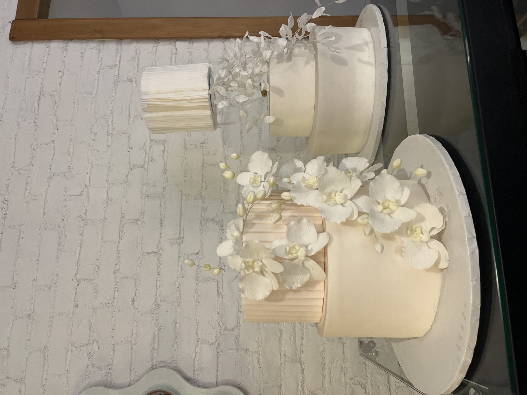 Two tiered wedding cakes are displayed on a wall. The top cake has a white finish and is decorated with white foliage. The bottom cake is a combination of horizontal lines and a smooth white finish, decorated with white orchids. The cakes are affixed to the wall, which is a white brick design. The environment suggests a showroom or shop, possibly in Varna, Bulgaria, though no definitive location is specified. There are no people visible.