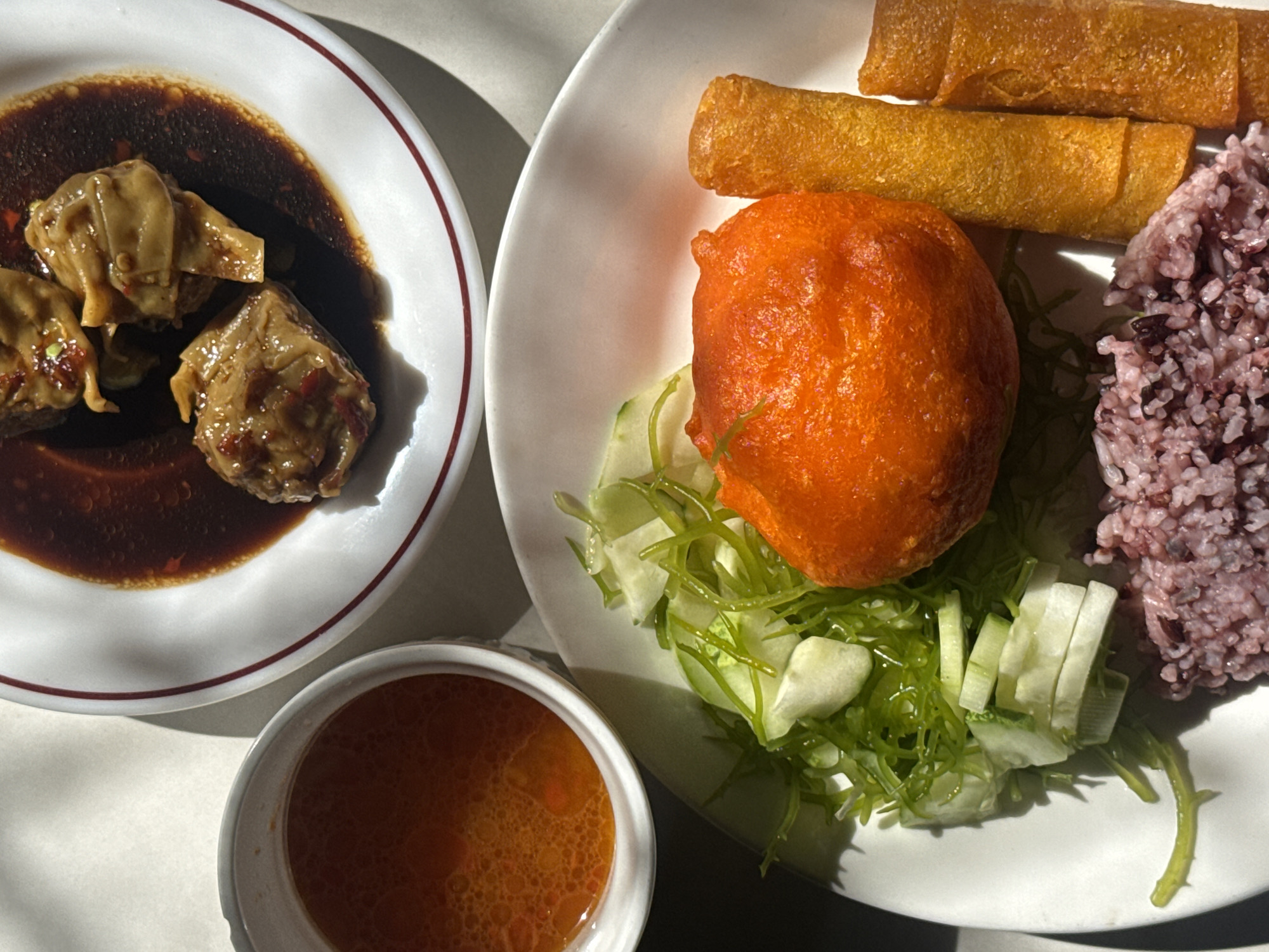 The image displays a meal served on a white surface, likely a table, with natural light casting shadows. In the foreground, a white plate holds a portion of purple rice, three golden-brown spring rolls, a bright orange, irregularly shaped food item, and a side salad composed of thinly sliced cucumbers and green, stringy vegetables. To the upper left, a small white bowl contains a reddish-orange broth with visible oil droplets. Behind these items, a larger white plate contains several pieces of dumplings bathed in a dark sauce, with some appearing slightly textured and others smooth. The composition suggests a culinary setting, potentially a restaurant or home dining environment in Davao City, Philippines, given the context provided. There are no people or animals visible in the image. The focus is entirely on the food and its presentation.